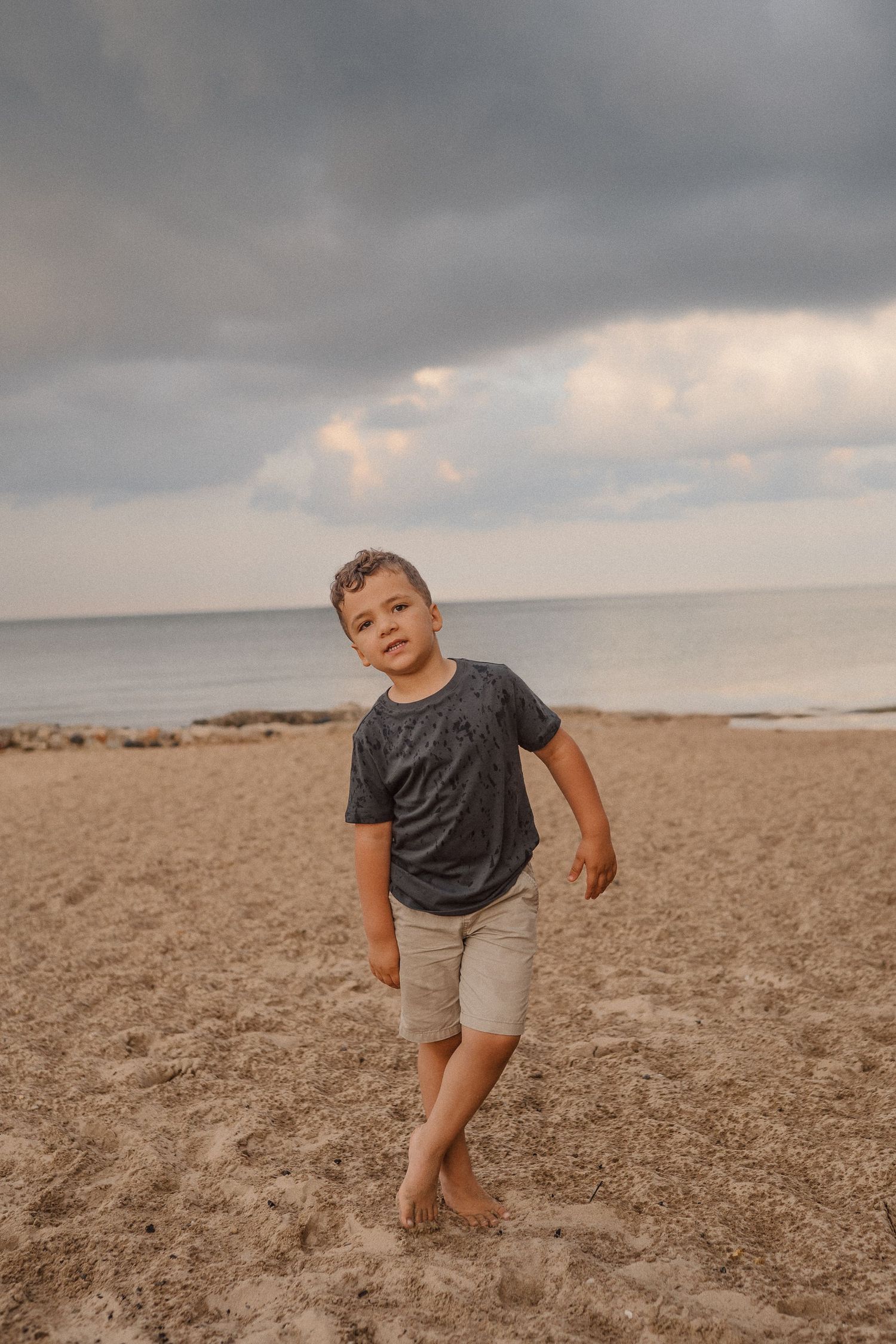 A young child plays joyfully on a sandy beach with dramatic storm clouds in the background.
