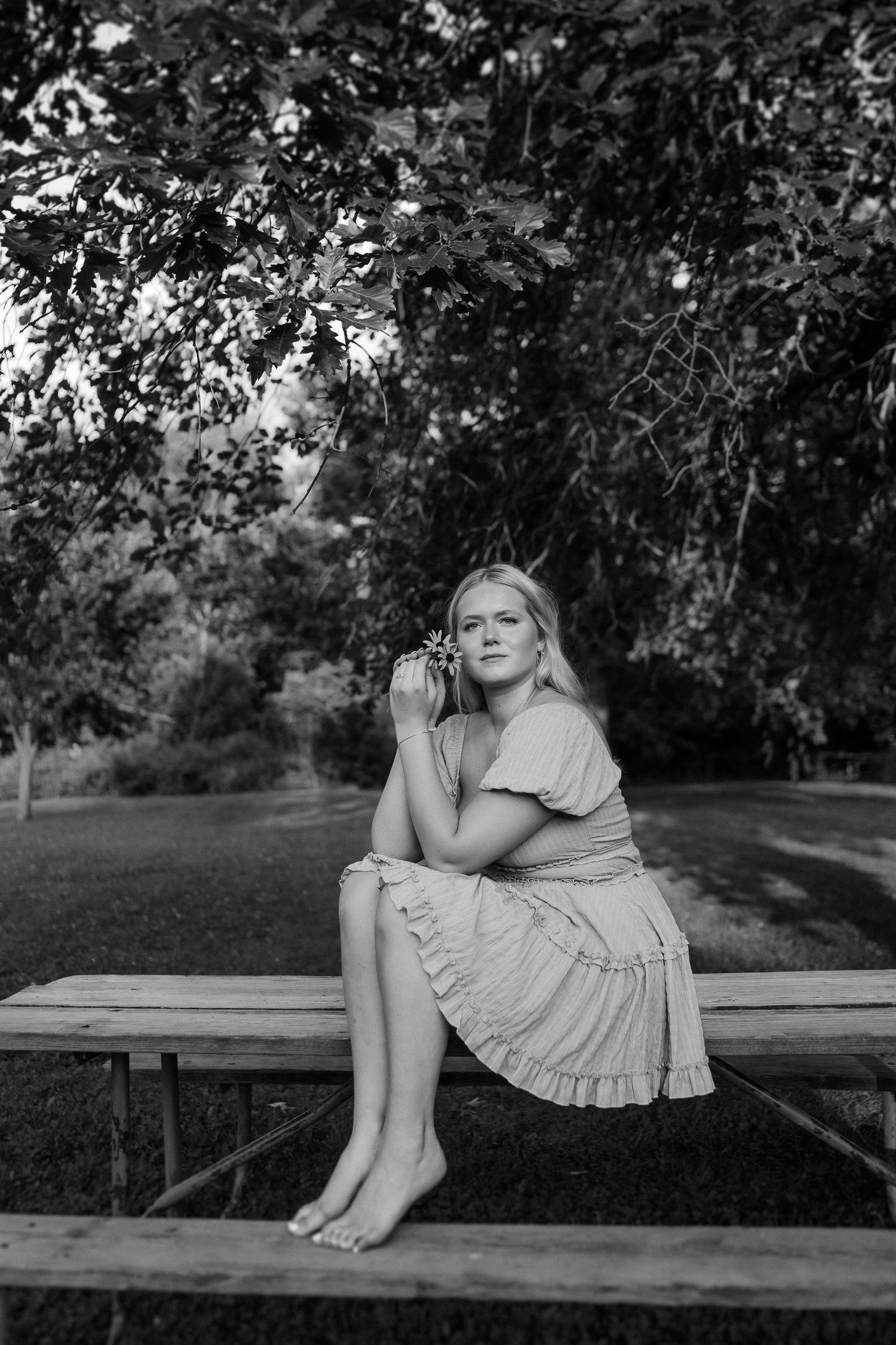 Person in a light-colored dress poses gracefully on a wooden bench in a natural outdoor setting.