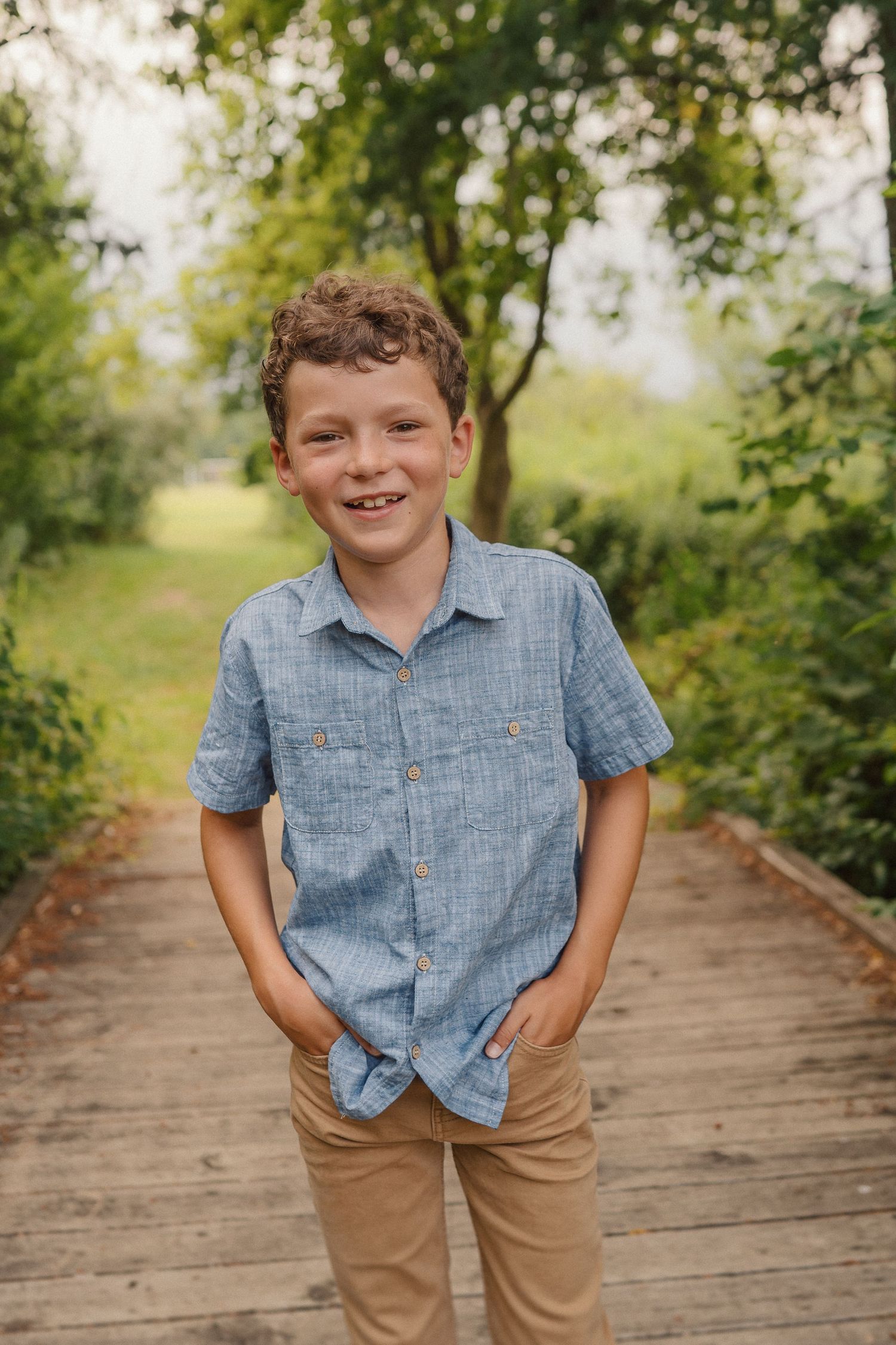 A young boy in a blue button-down shirt stands on a wooden walkway surrounded by greenery.