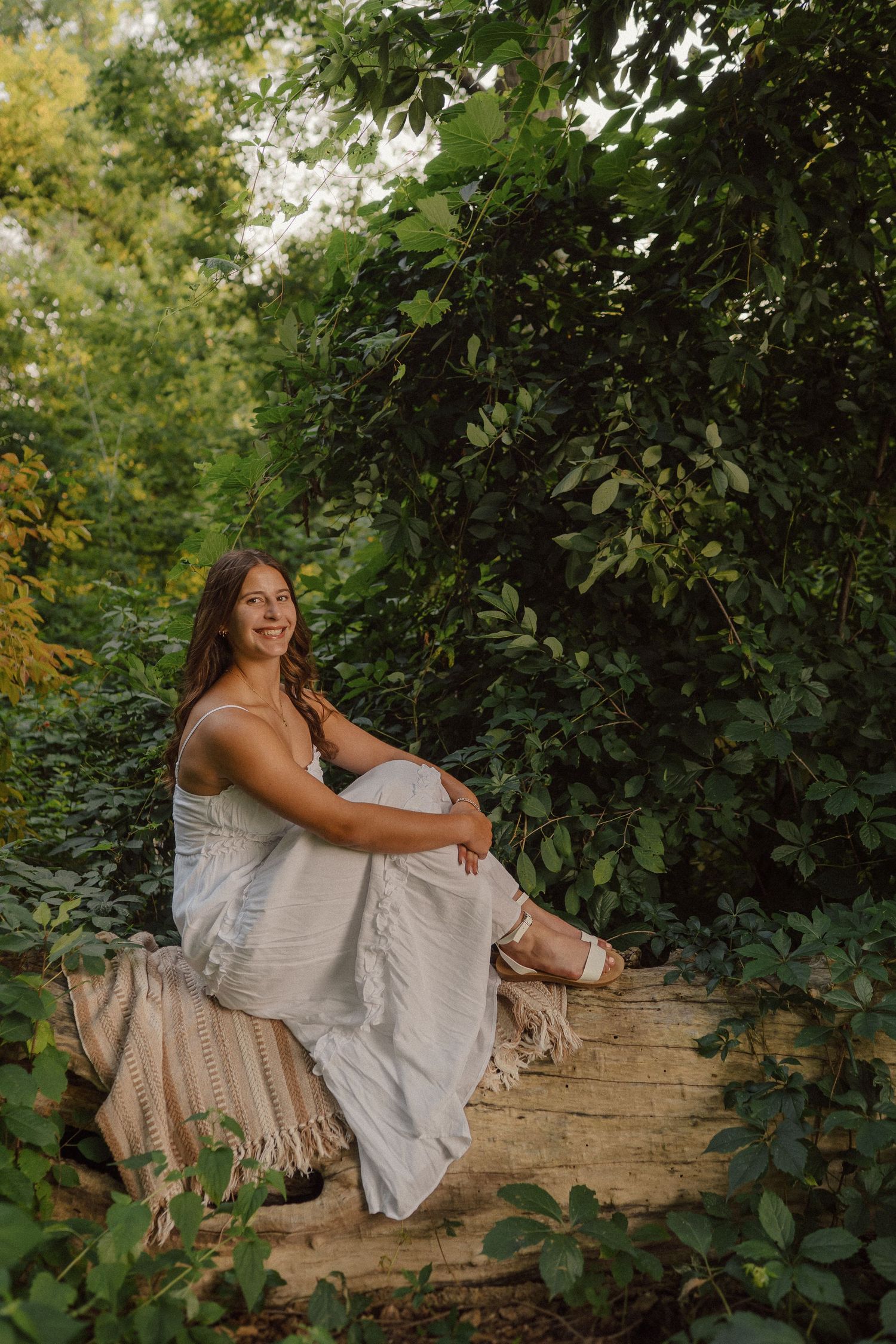 A person in a white summer dress poses on a wooden log surrounded by lush green foliage on a sunny day.