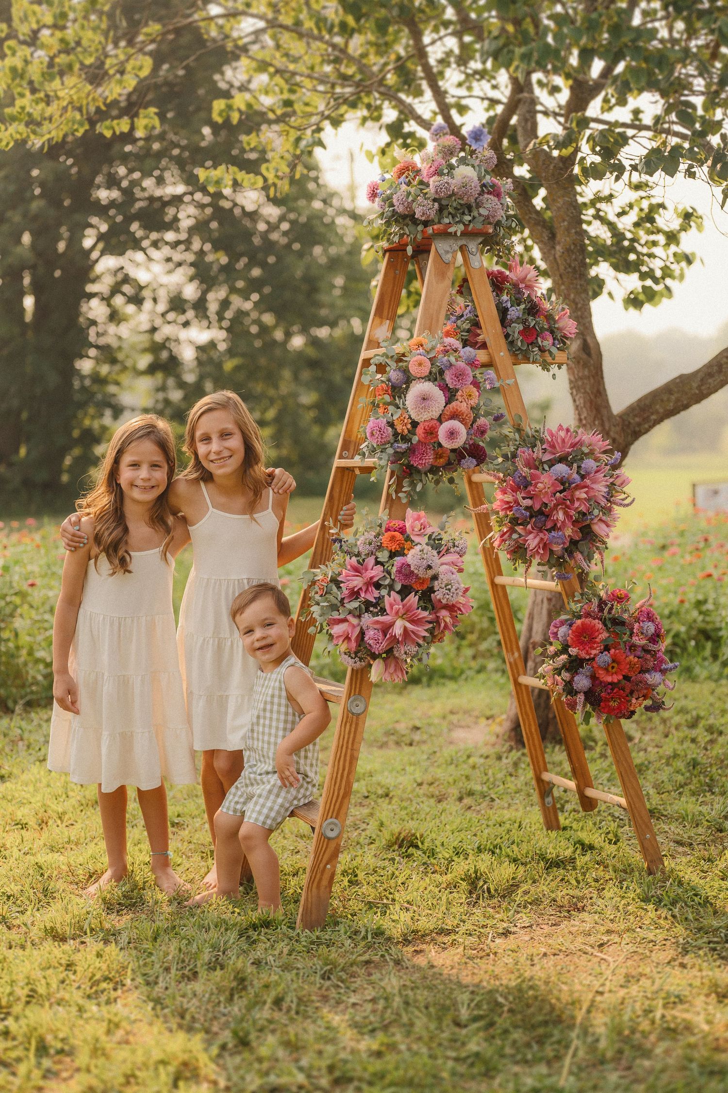 Children in white dresses pose near a wooden arch decorated with pink flowers in a sunny garden.