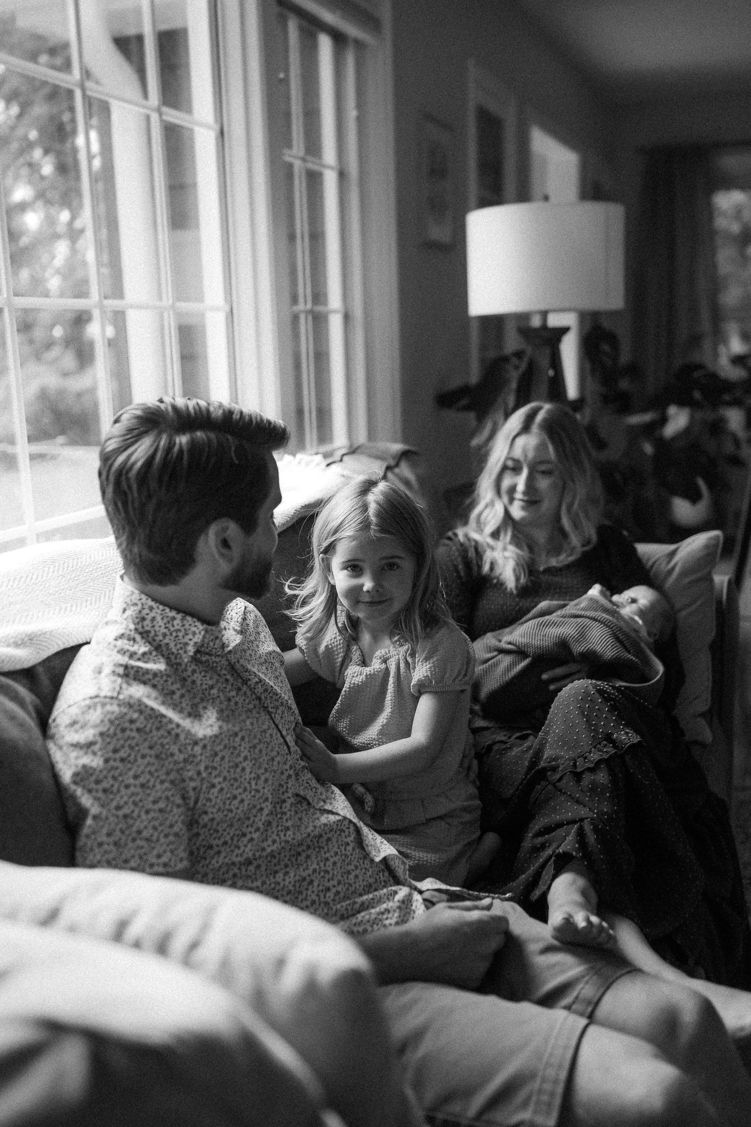 Black and white photo of family sharing an intimate moment together while relaxing on cozy furniture near window.