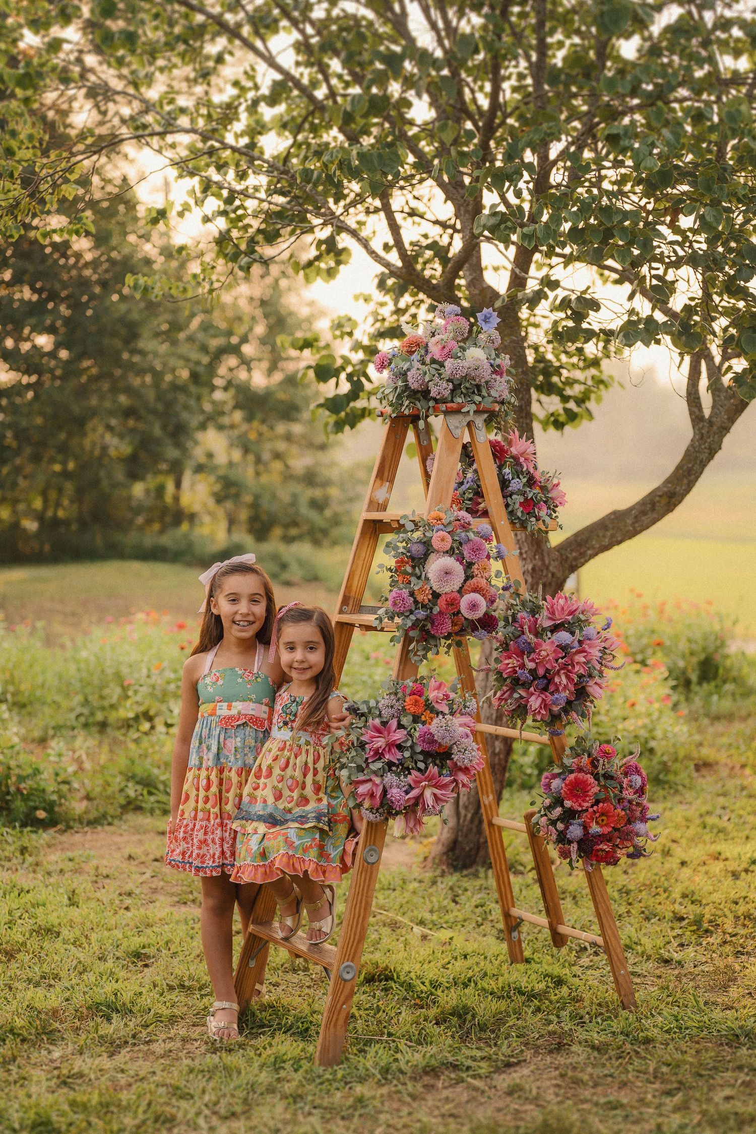 Kids in colorful summer dresses pose next to wooden easel with bright pink floral arrangement in sunlit garden setting.