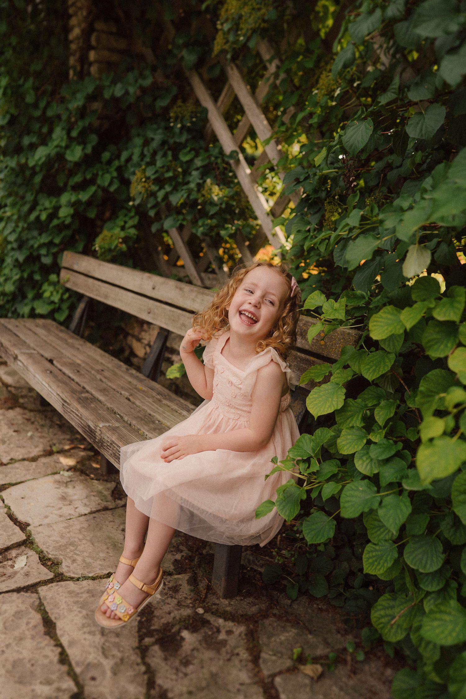 A child in a pink dress sits on a rustic wooden bench surrounded by lush garden greenery.