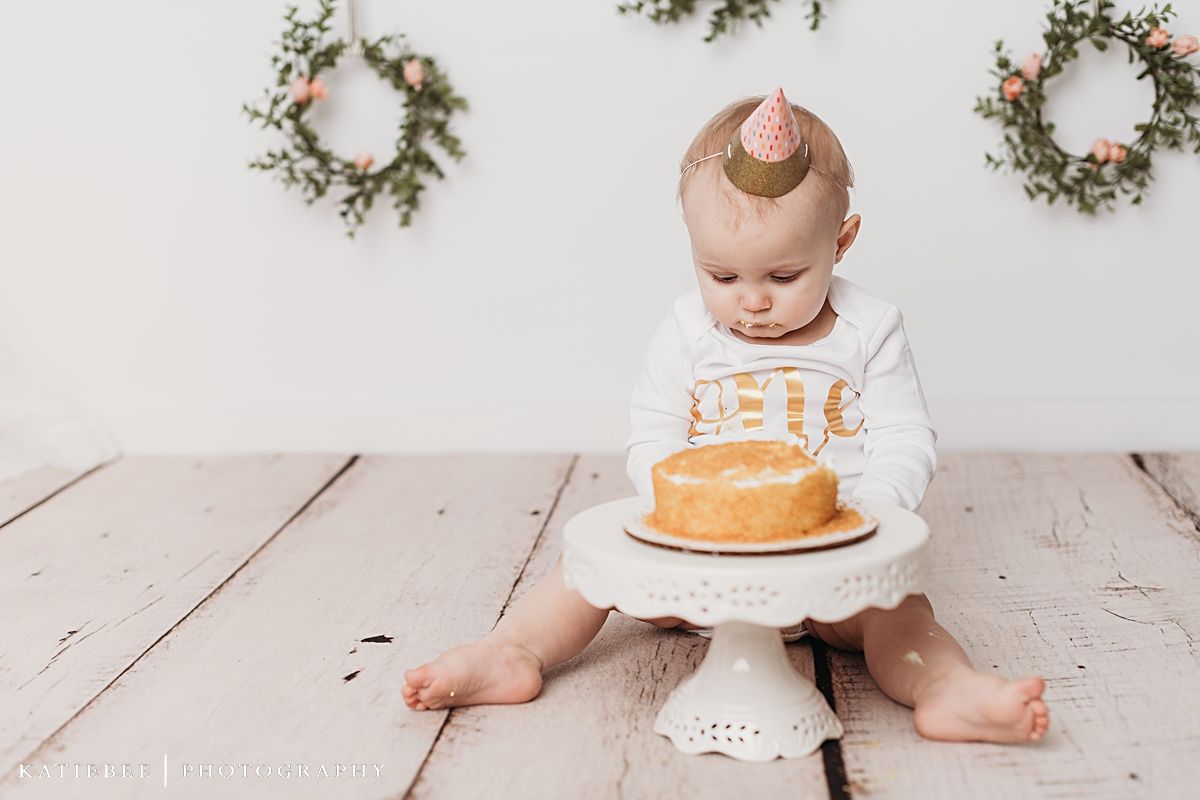 Baby in festive attire expressing excitement near cake with decorative wall wreaths.