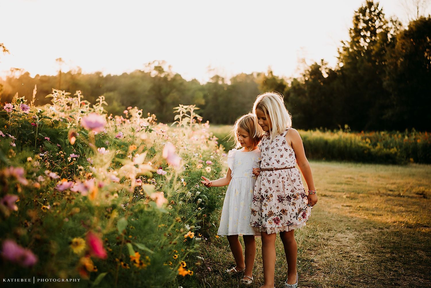 Two figures stroll along a garden path at golden hour surrounded by blooming flowers.