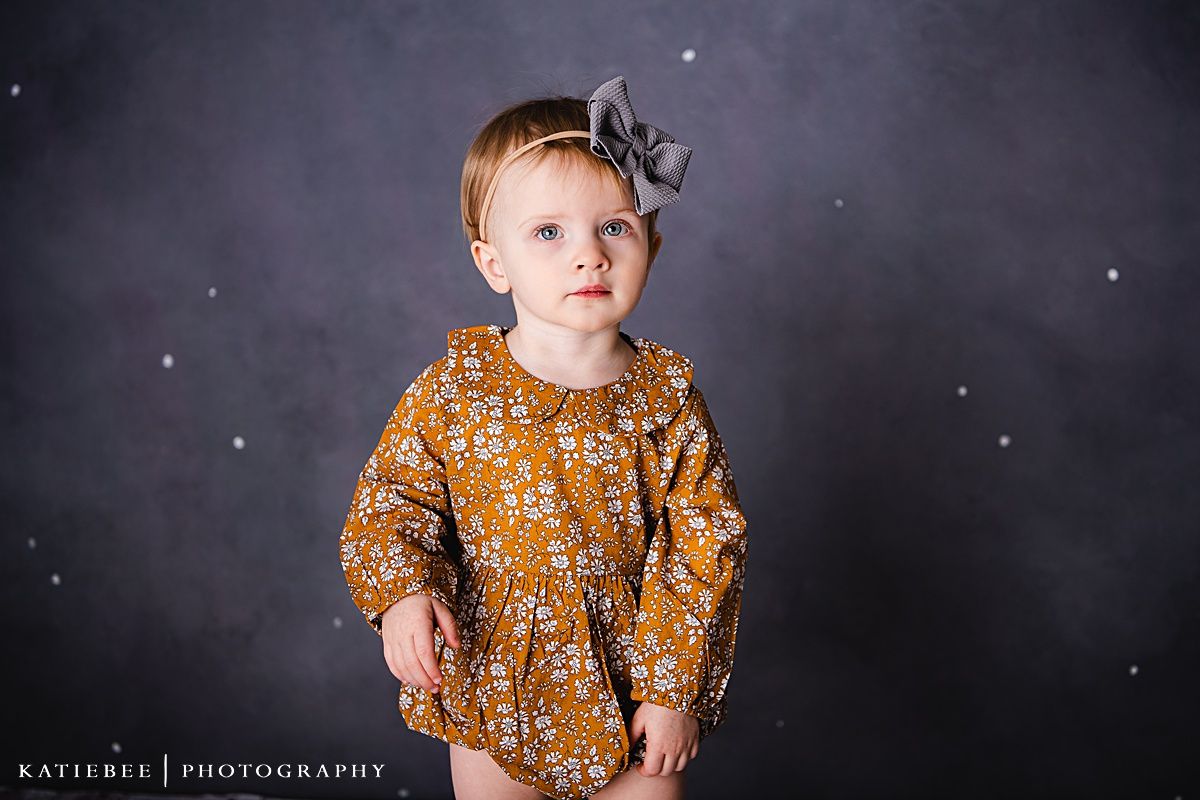 A toddler wearing a mustard floral dress and grey hair bow stands against a dark backdrop.