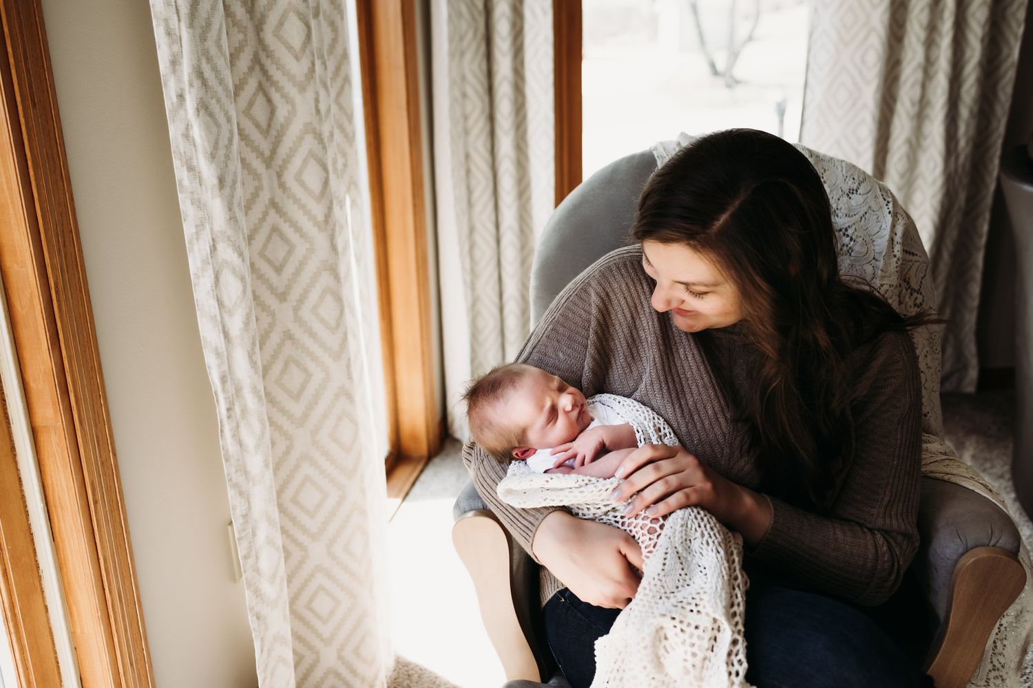 A tender moment by a window as a baby wrapped in knit blanket is cradled in brown sweater-clad arms.