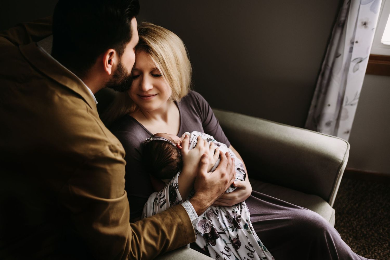 Intimate indoor portrait of couple on grey couch holding newborn baby.
