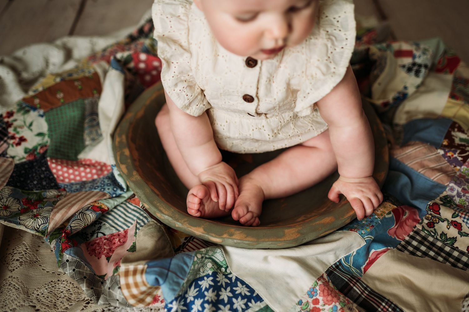Baby in cream romper sits on wooden bowl surrounded by colorful vintage quilt pieces.