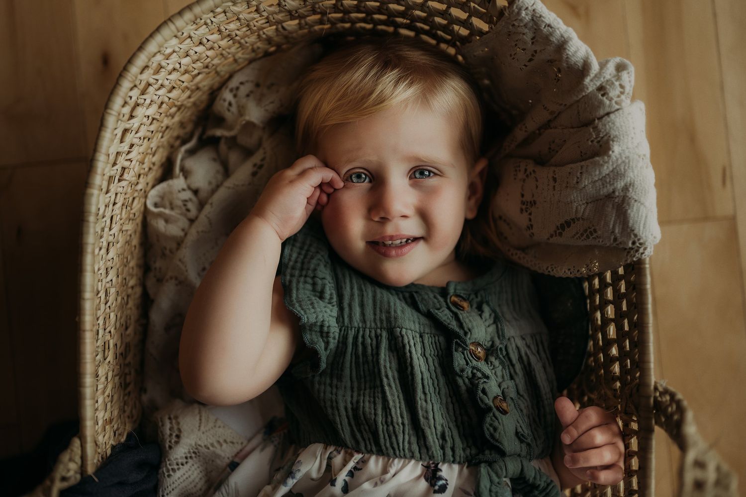 Toddler wearing green dress sits in woven basket chair with joyful expression.