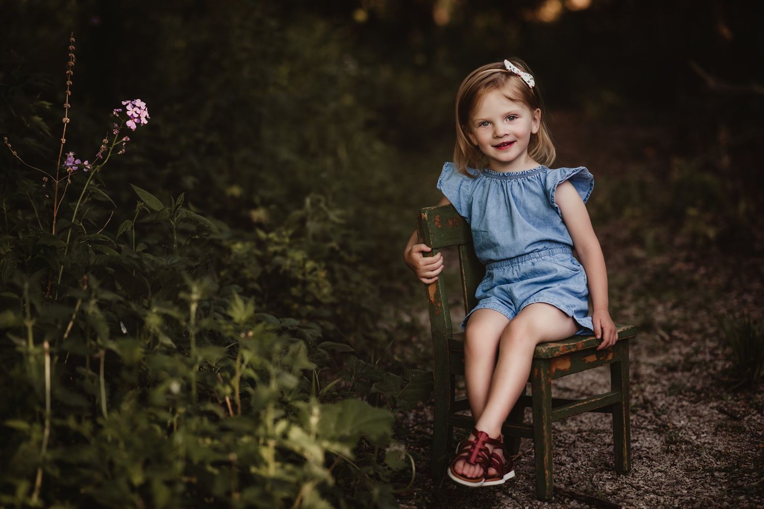 Child in blue romper sitting on wooden chair outdoors near purple wildflowers.