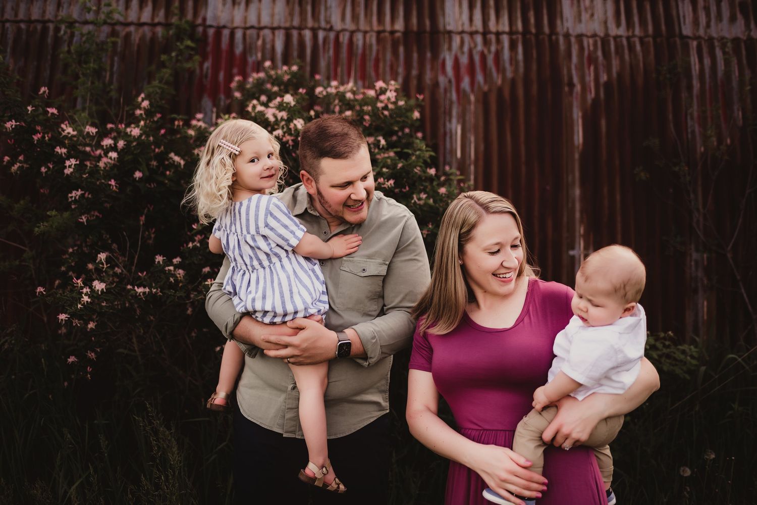 Casual family photo session against a weathered barn with blooming flowers.