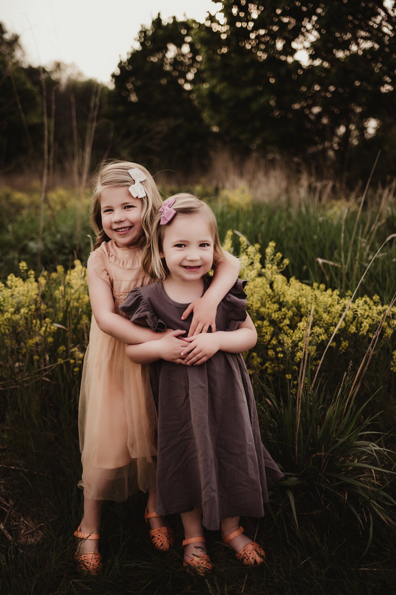 Two children in dresses share embrace while standing among yellow wildflowers at dusk.