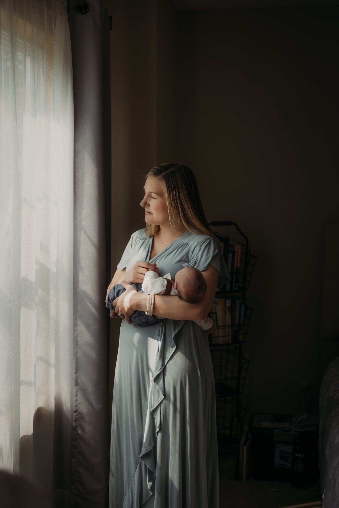 Parent wearing blue dress holds sleeping infant by window in dimly lit room.