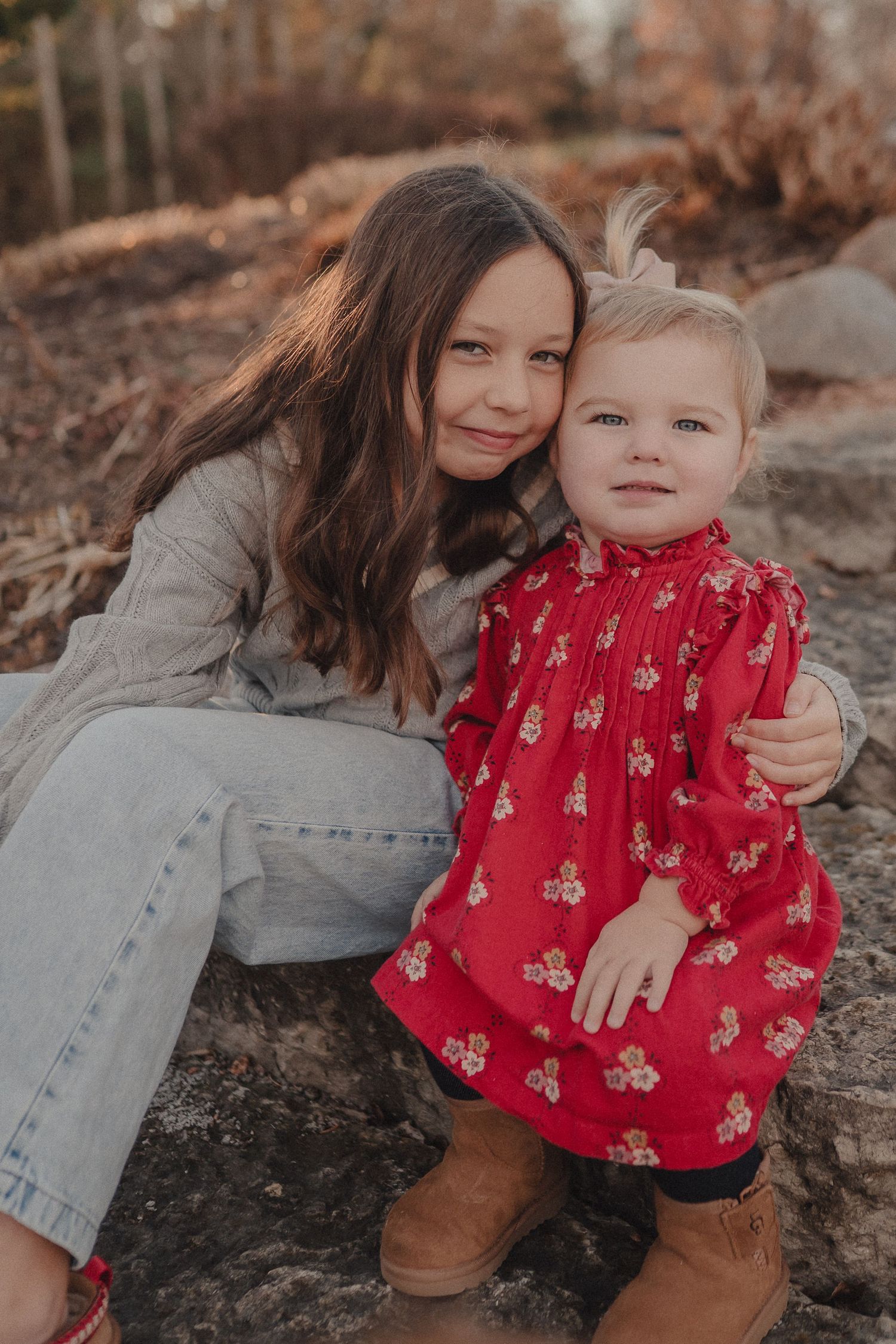 Two people sit together on rocks outdoors, one wearing a red floral dress and one in casual denim.