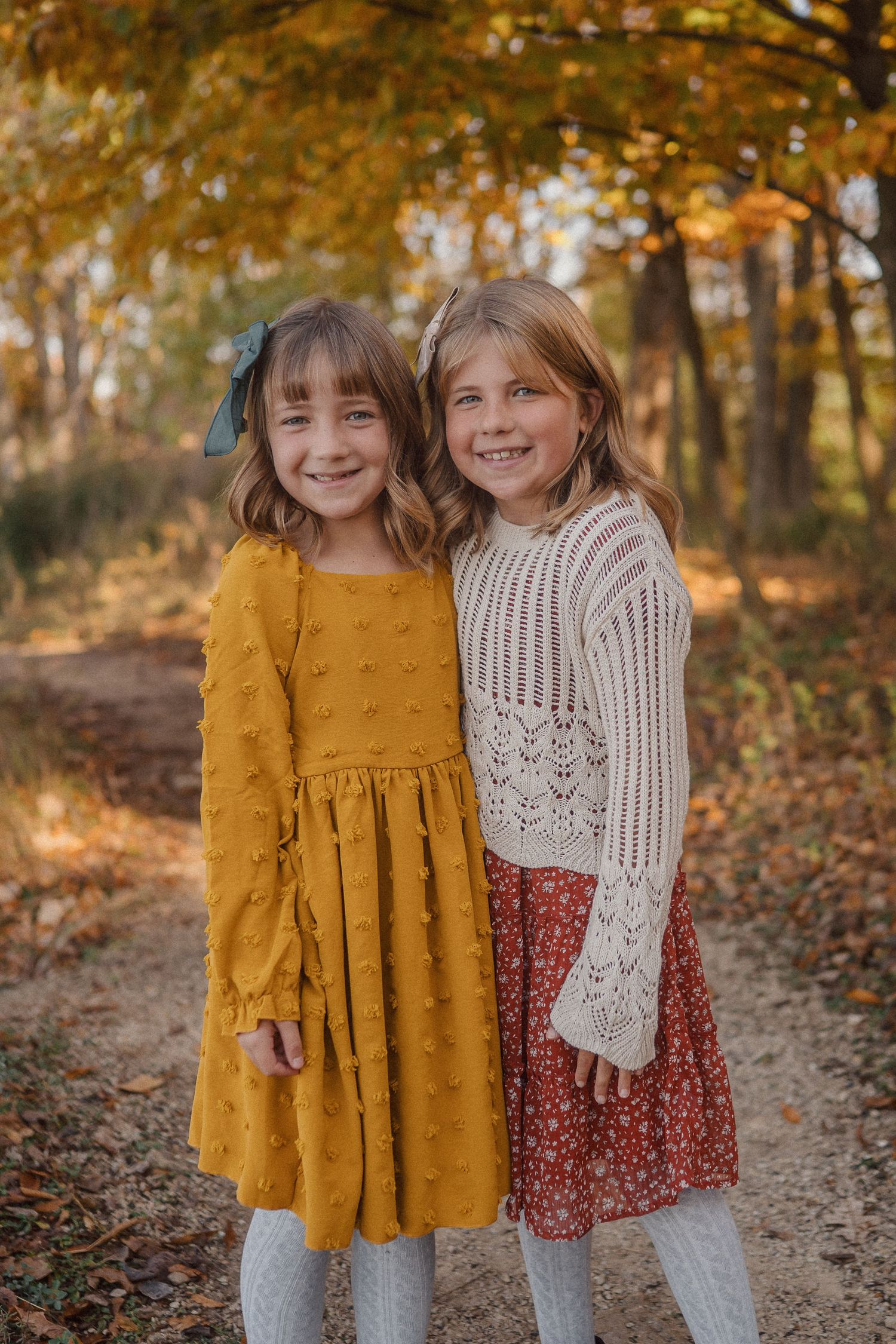 Two children in fall outfits standing together on a leaf-covered path with autumn trees in the background.