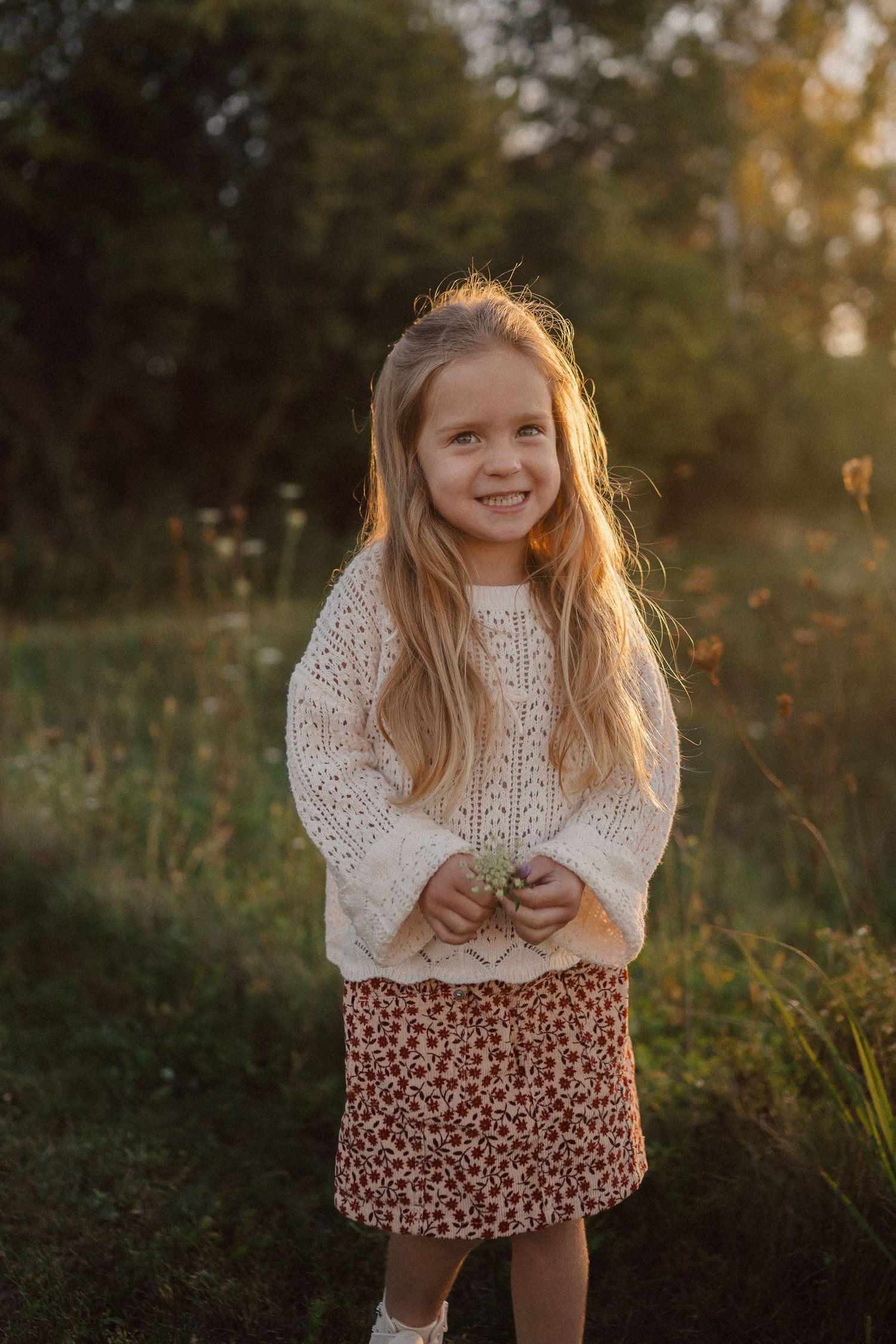 Young girl in white sweater and floral skirt smiles during golden hour photoshoot in nature.