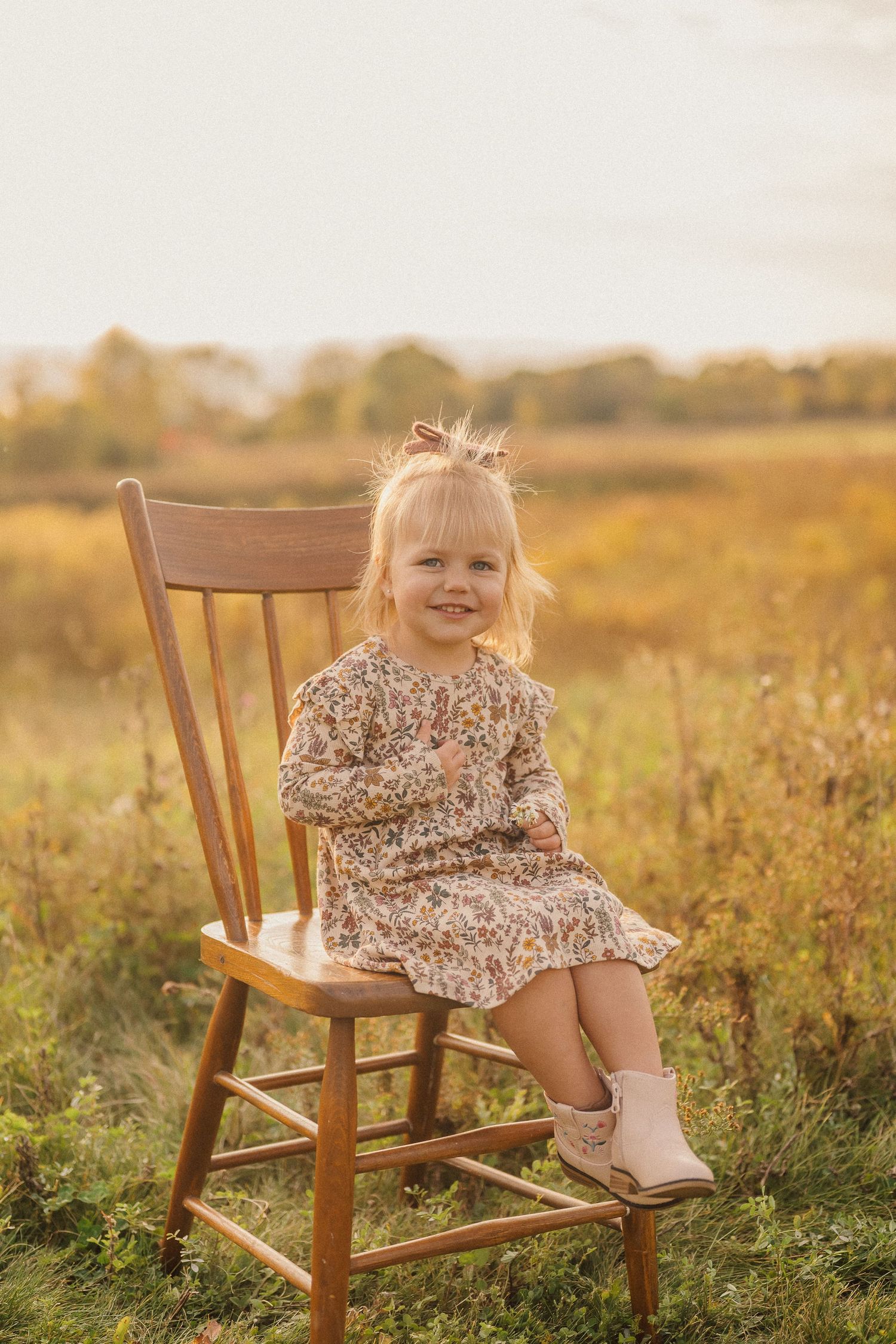 Small child in floral dress sitting on wooden chair in sunlit meadow at golden hour.