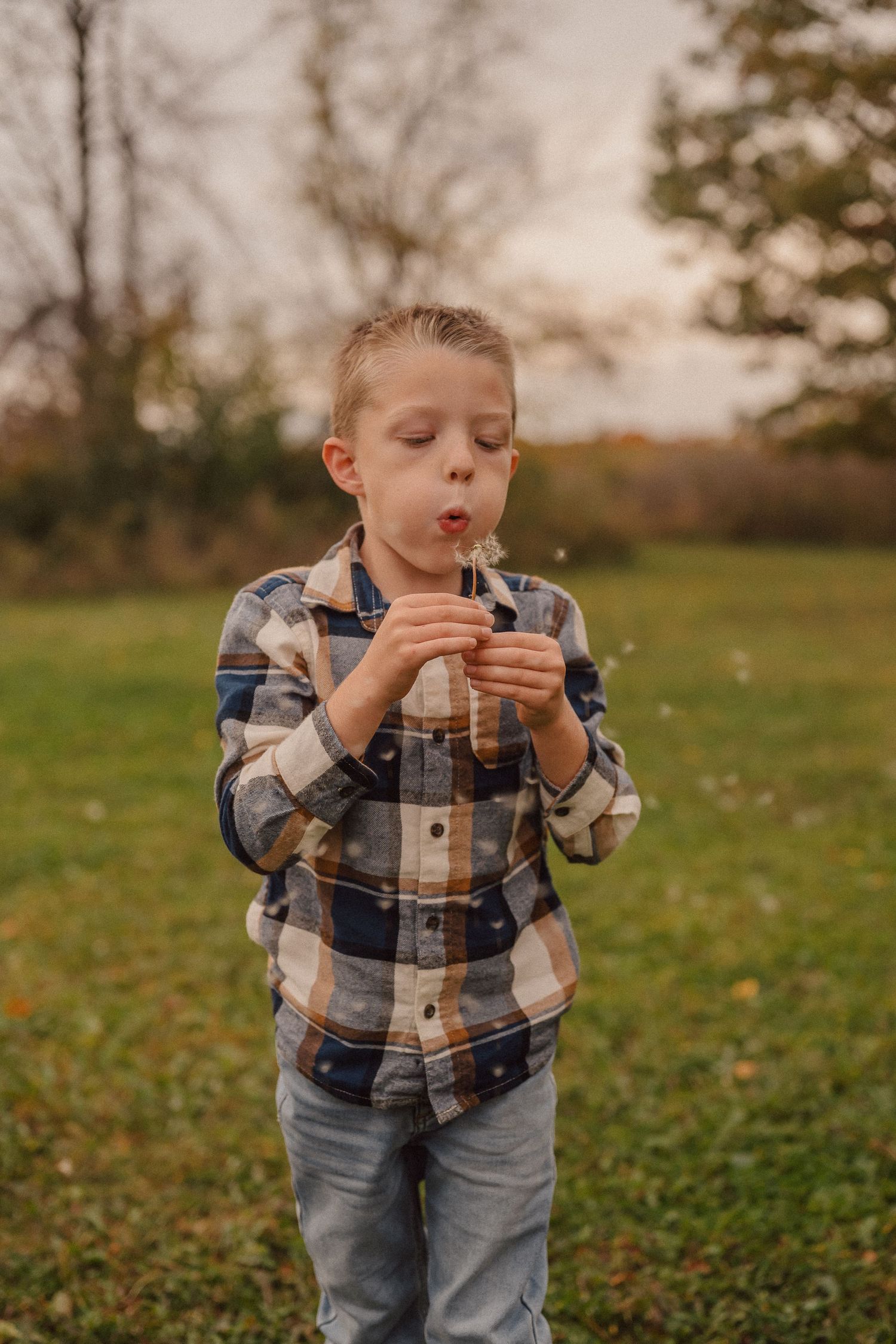 A child in a plaid shirt blows on a dandelion in a grassy field during fall.