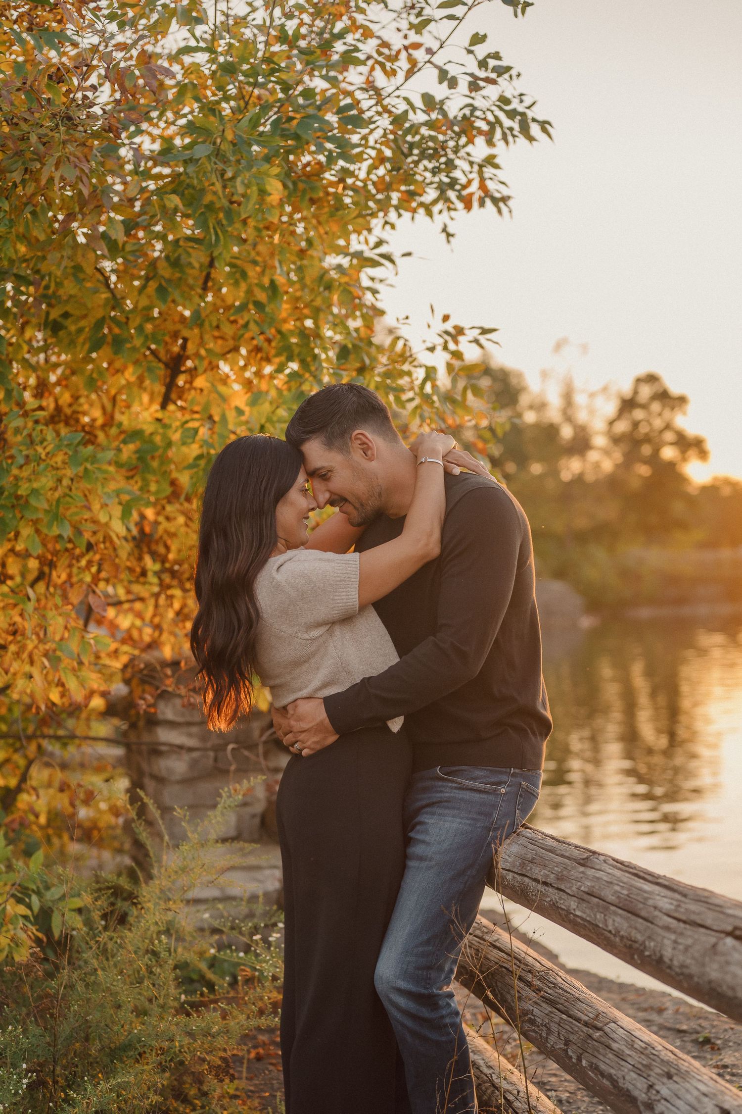A romantic couple embracing by a lake during golden hour with autumn foliage in the background.