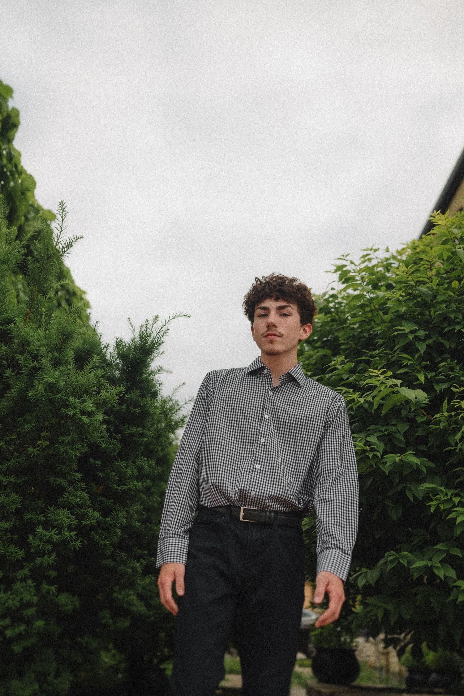 A casual portrait shot of someone in a gingham shirt and dark pants standing between green hedges.