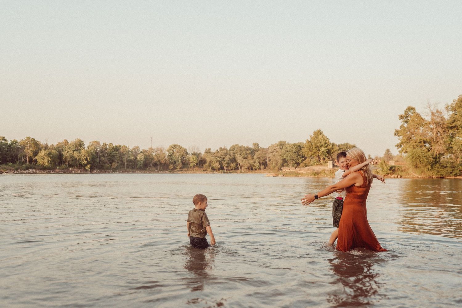 A couple wades with their child in calm lake waters at sunset surrounded by trees.