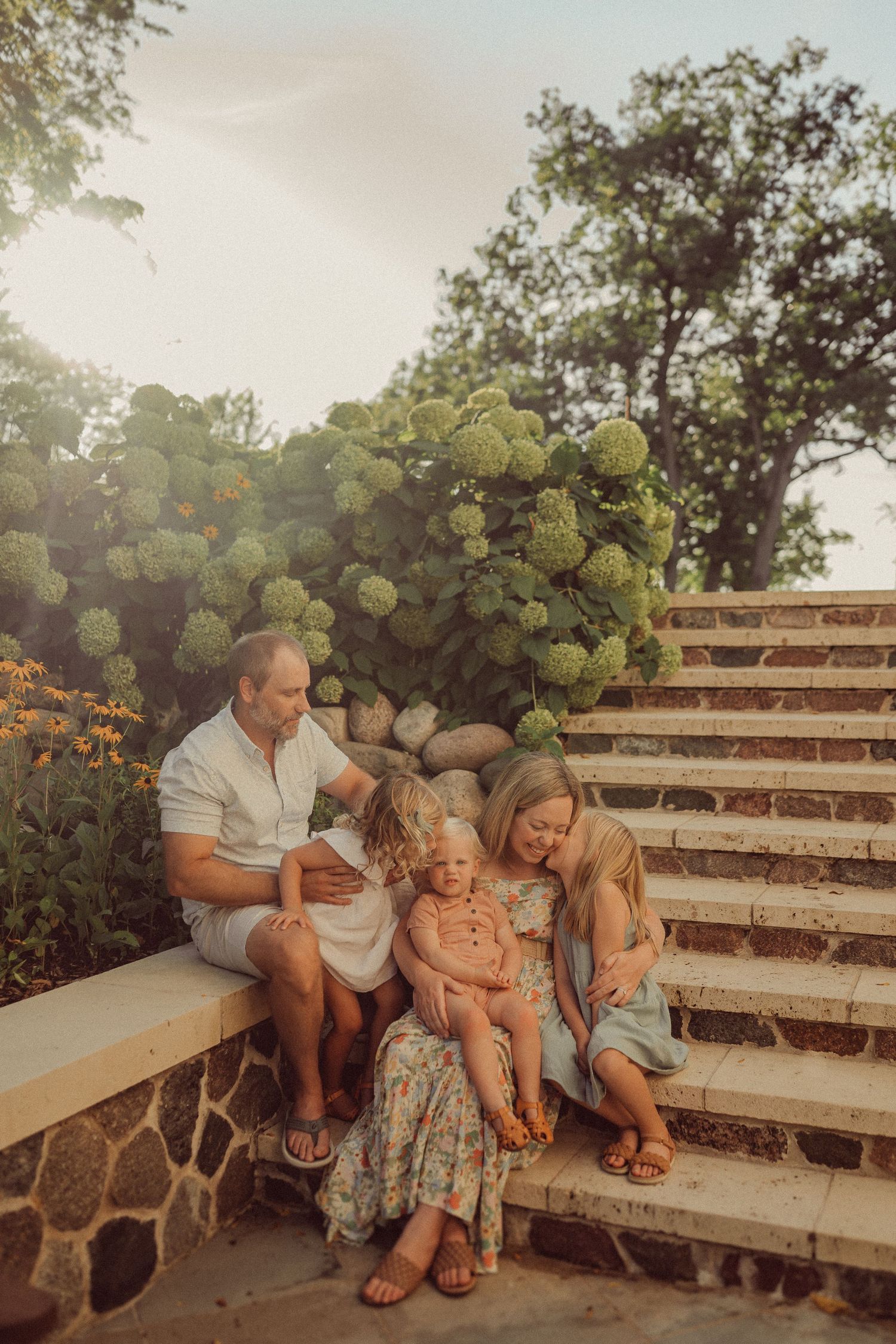 Family sitting together on rustic stone steps with hydrangea bushes in golden evening light.