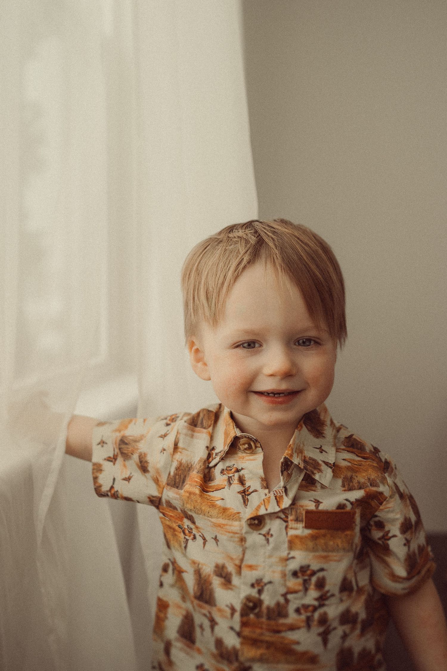 Close-up portrait of smiling toddler in brown patterned shirt against white wall.