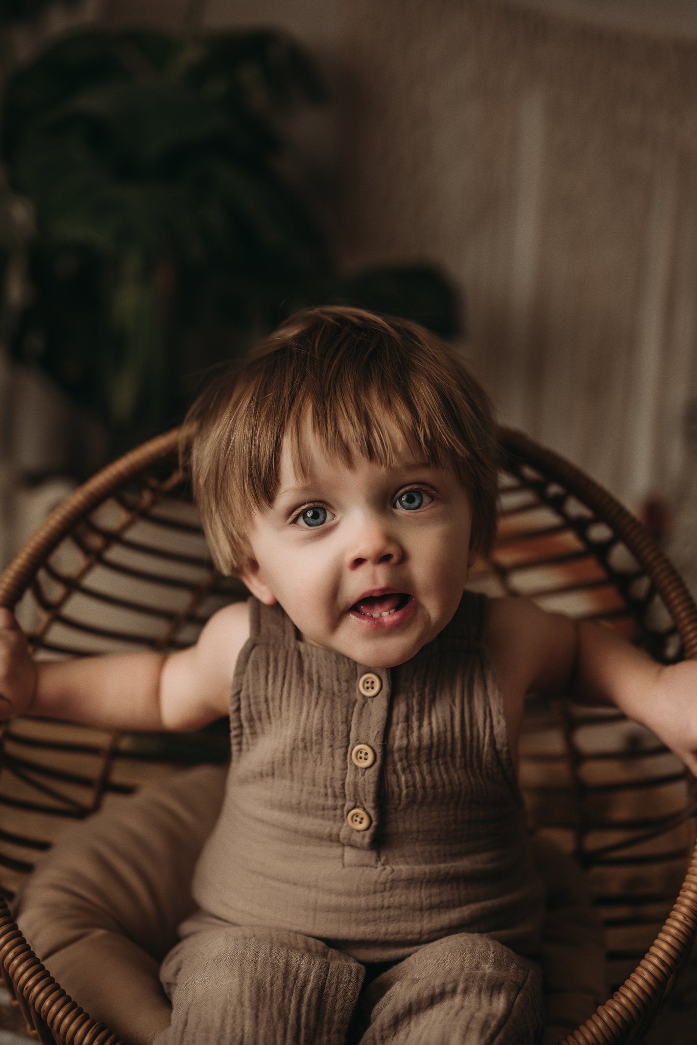 A toddler in tan clothing sits in a wicker chair against a boho-styled backdrop with plants.