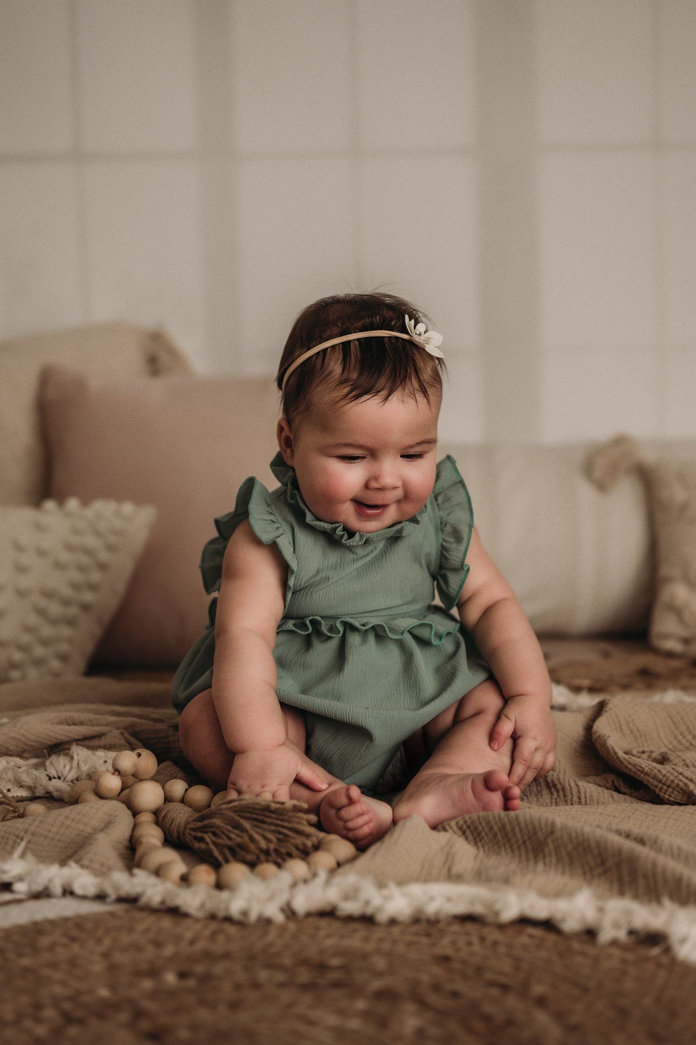 A baby in sage green ruffled dress sits on textured blankets with decorative wooden beads.