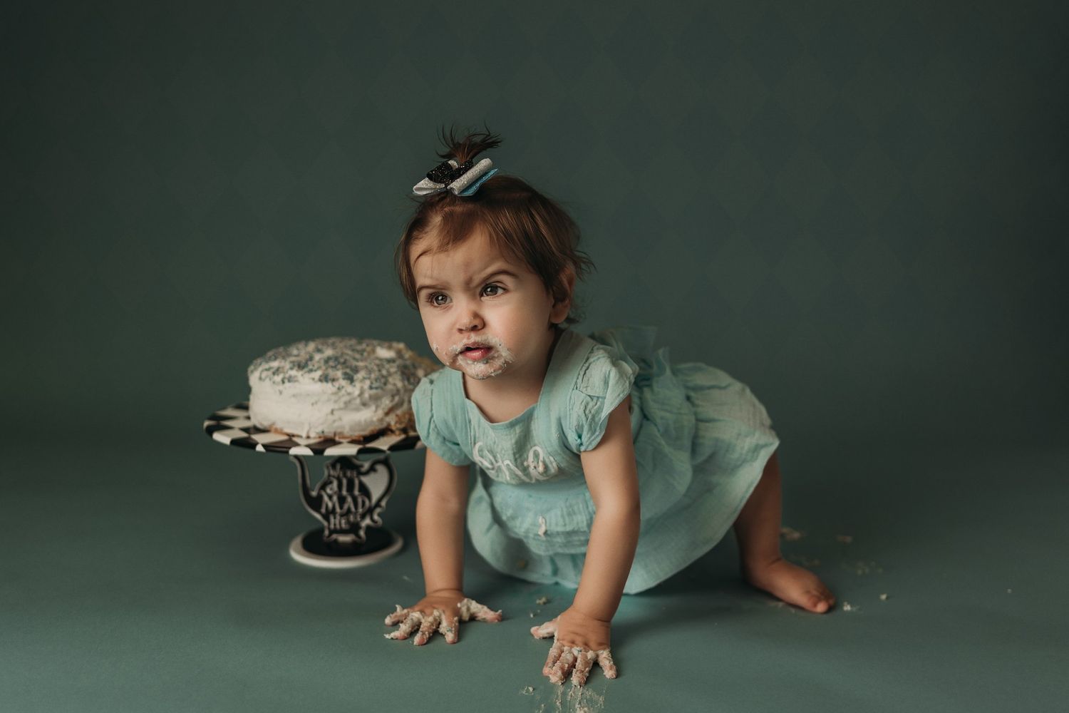 A small figure in a ruffled mint dress crawls near a decorated cake on an elegant stand.