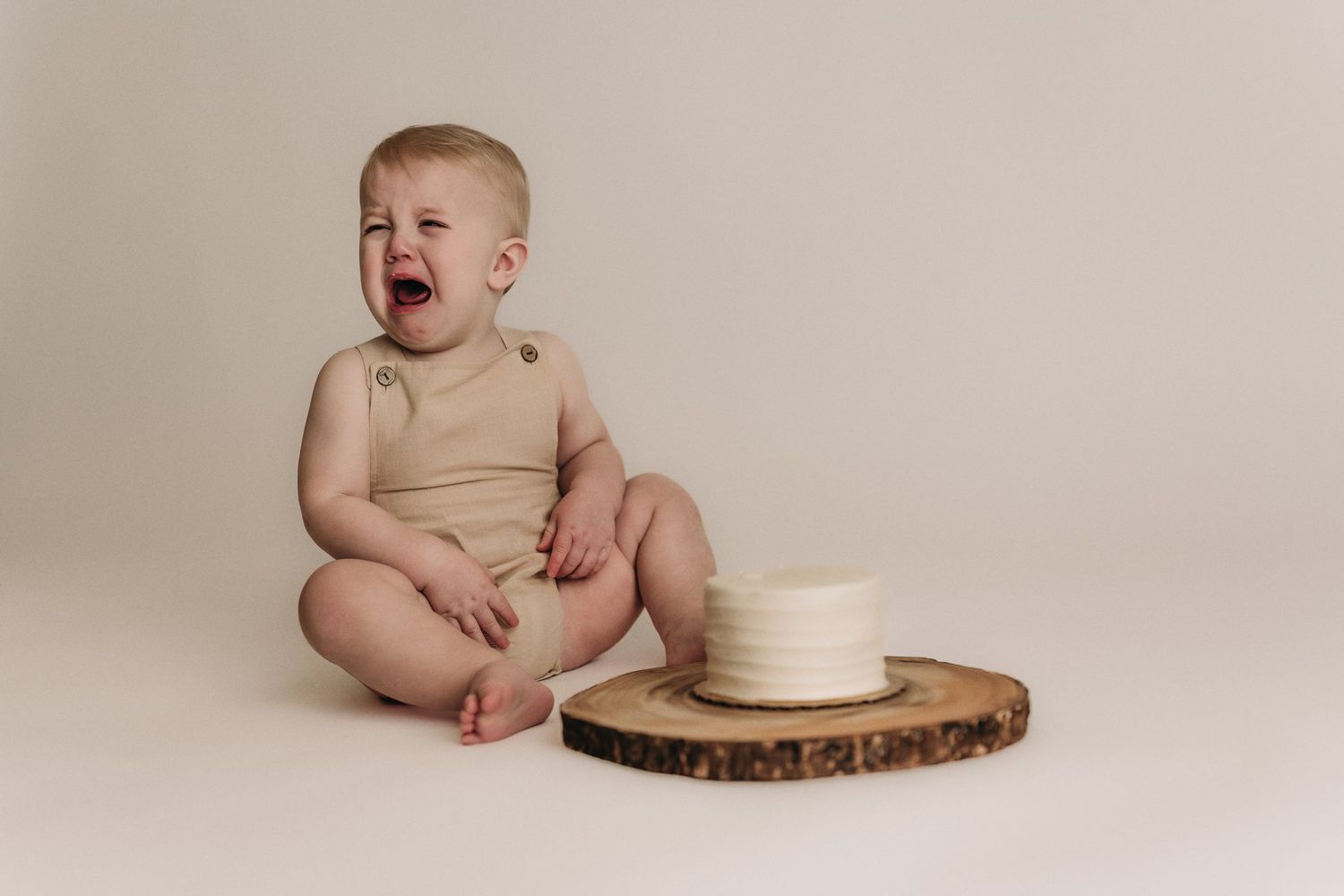 Baby in tan romper cries while sitting beside white cake on wooden board.