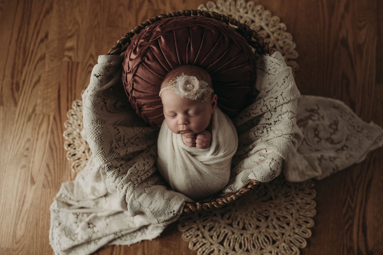 Overhead view of newborn in cream wrap nestled in round brown cushion with lace accents.