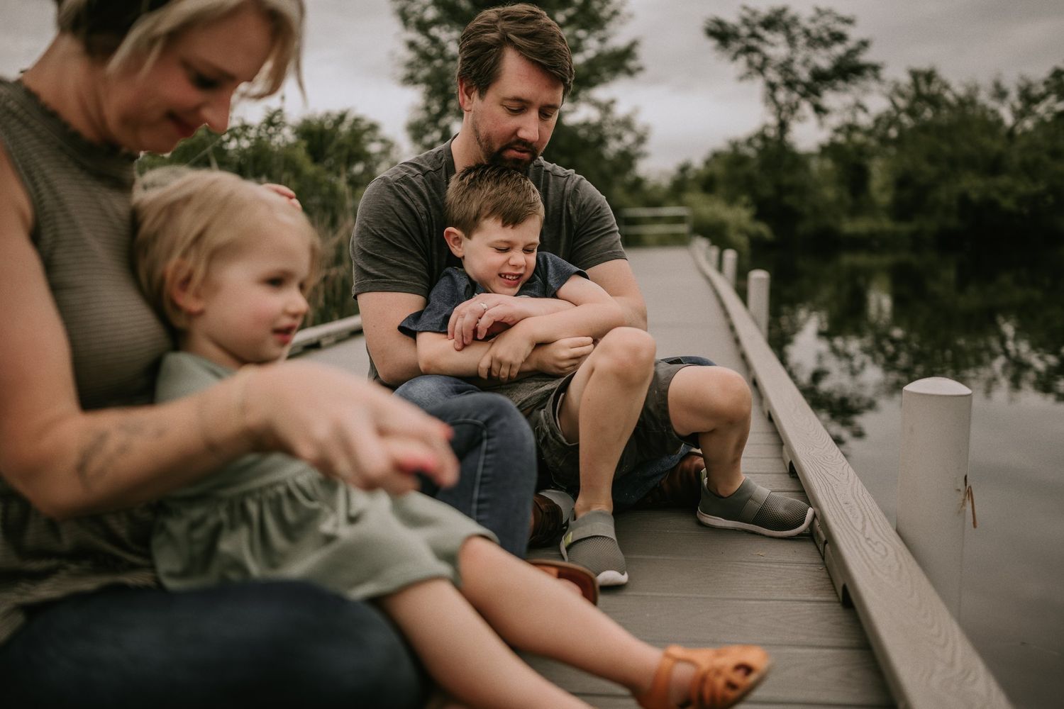 Family sits together on wooden dock overlooking tranquil pond at dusk.