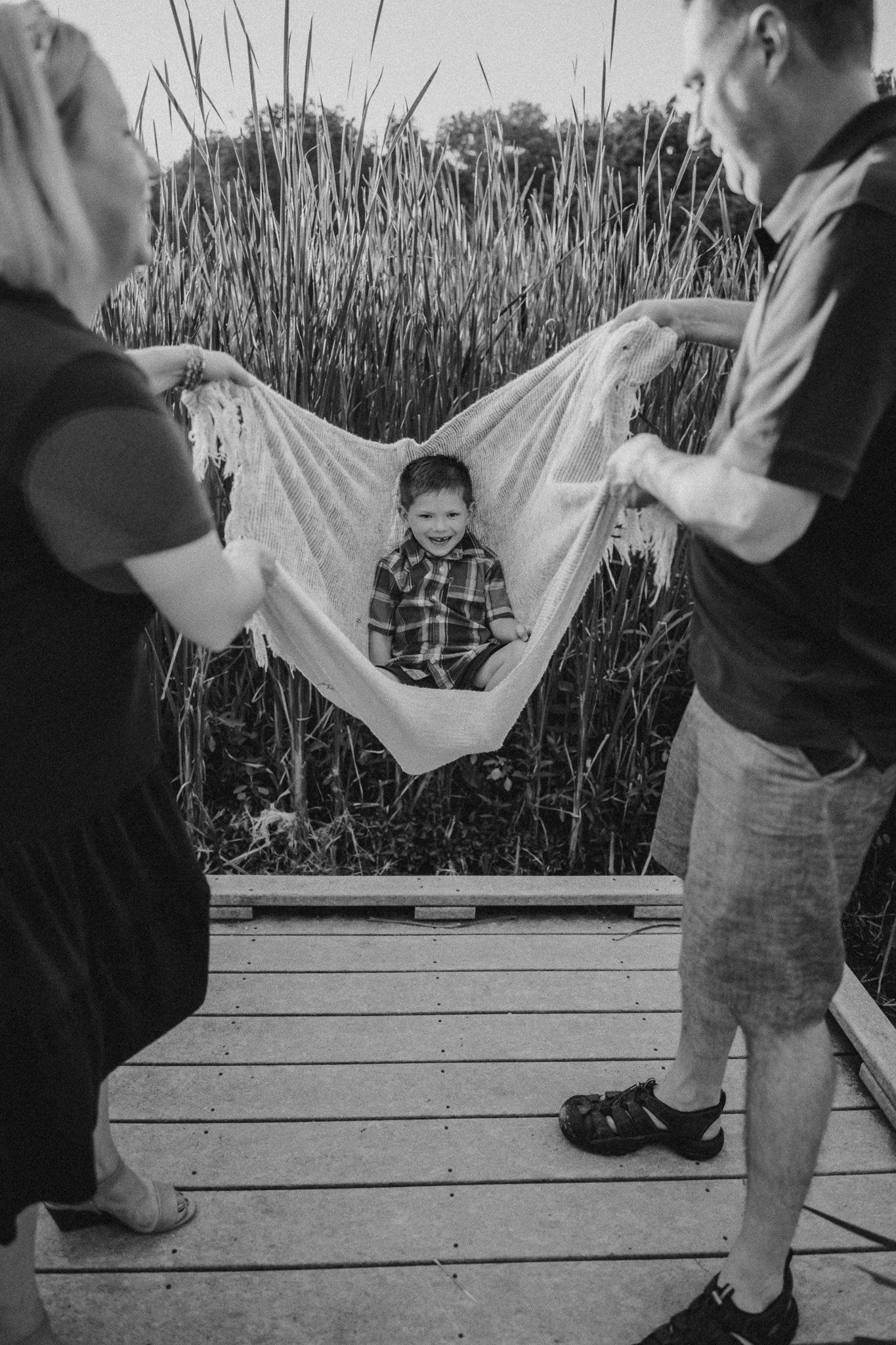 Child being lifted in a white blanket on wooden boardwalk surrounded by tall grass.