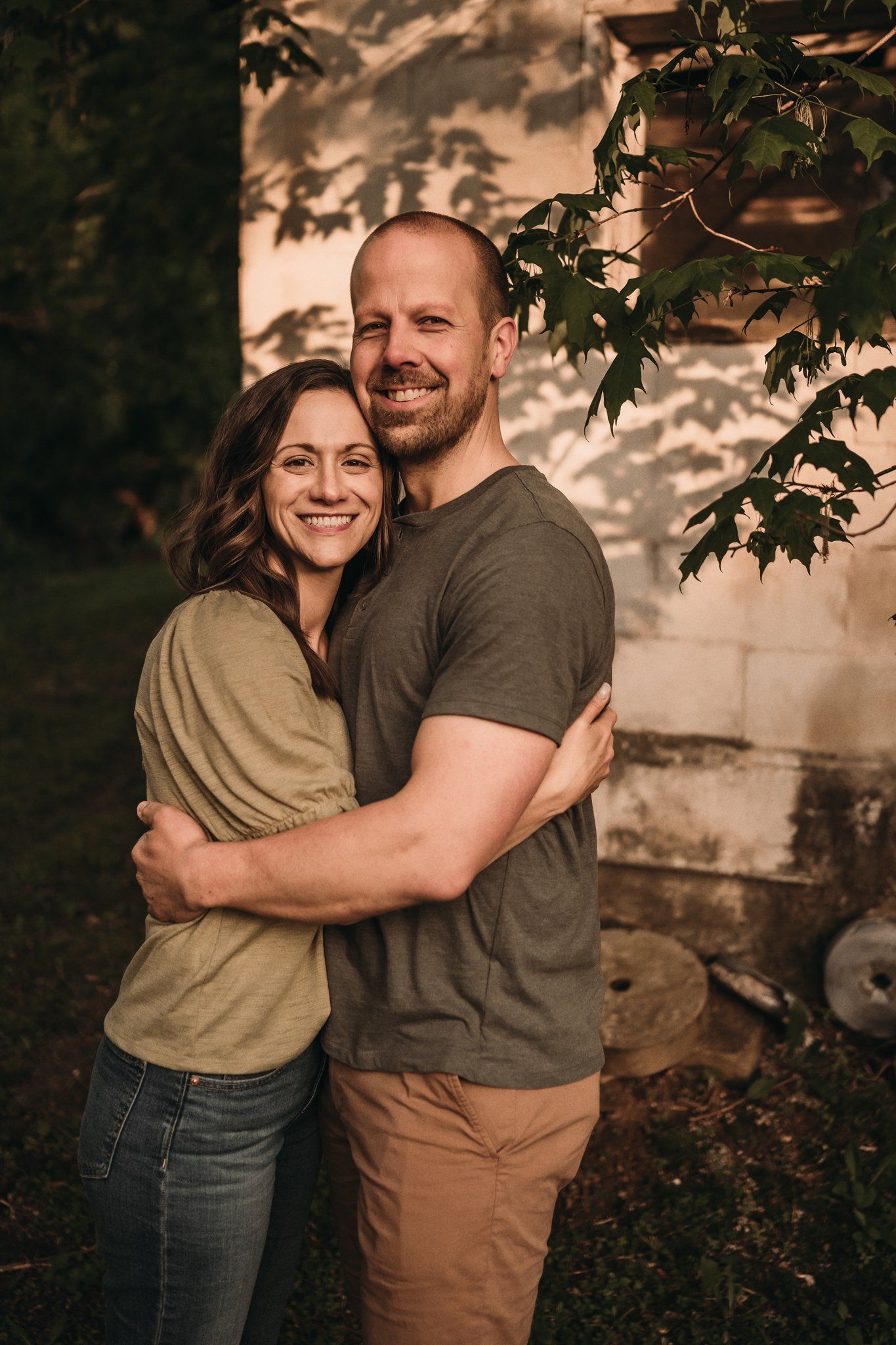 A couple embraces while smiling outdoors against a textured wall with leafy shadows.
