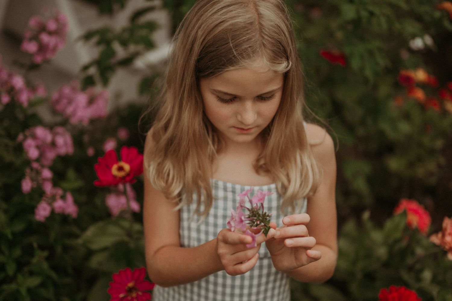 Young person in gingham dress examining flowers in a colorful garden setting.