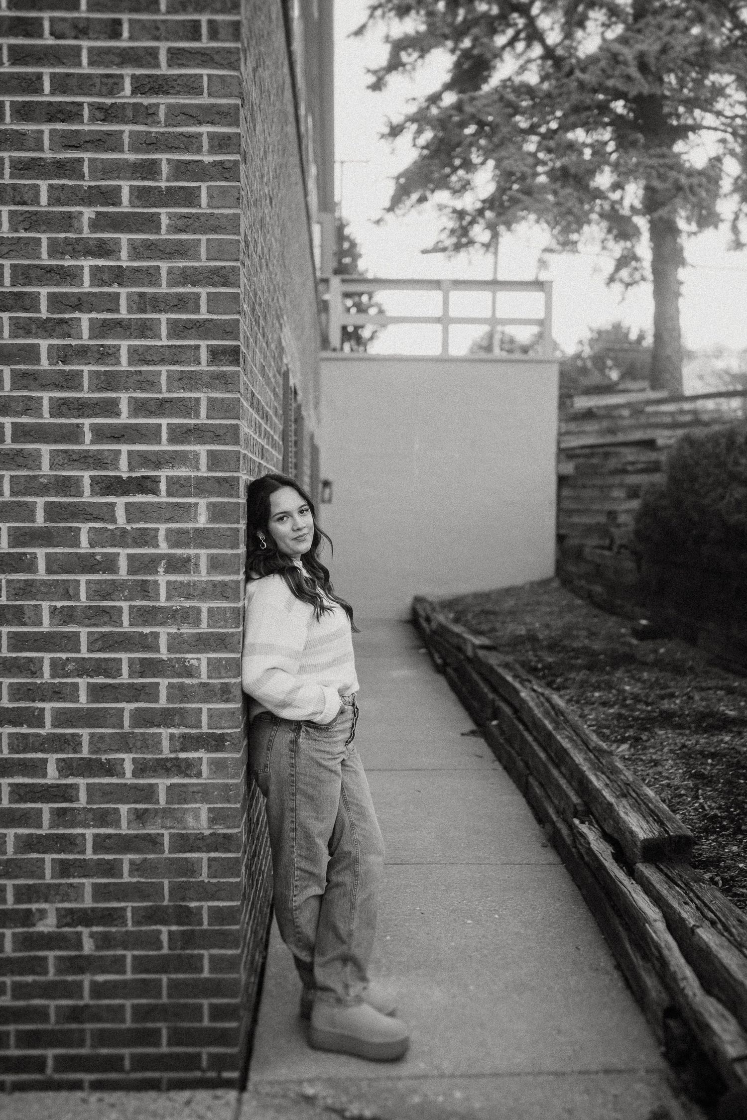 A casual portrait against a brick wall and concrete walkway in black and white.
