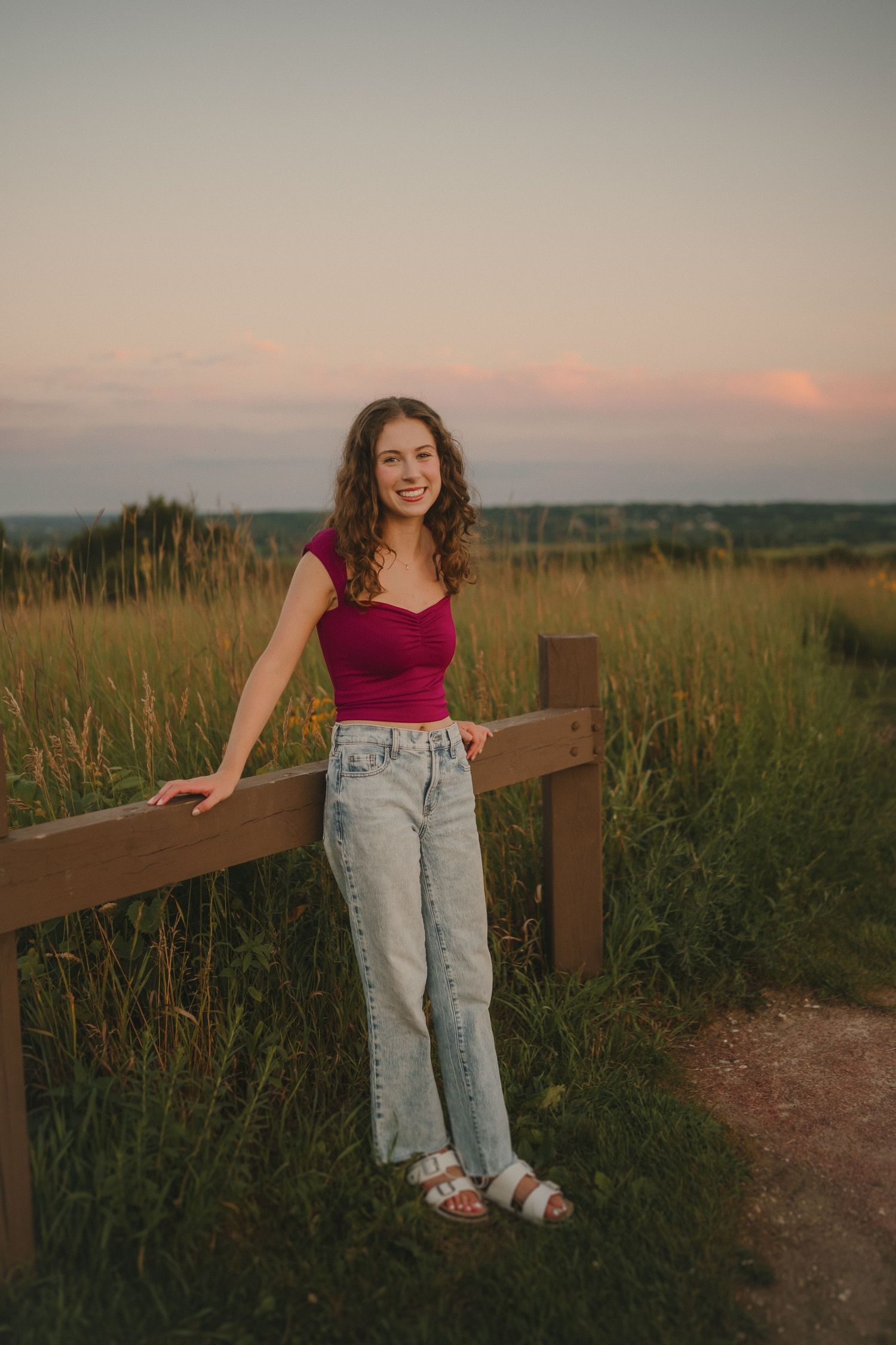 A person in a burgundy top and light jeans poses casually against a wooden fence during golden hour sunset.