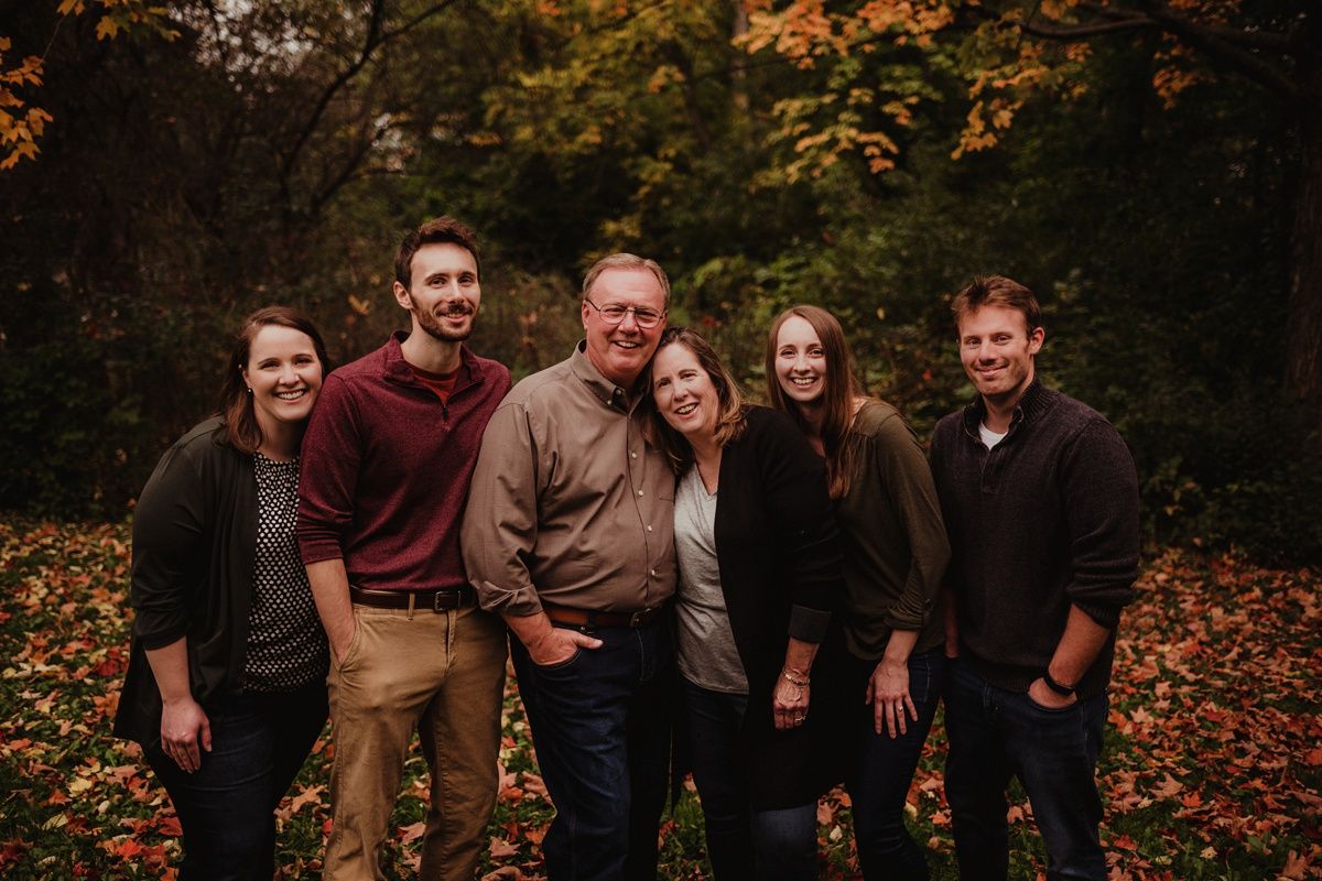 Family group photo of six people smiling together in autumn forest setting.