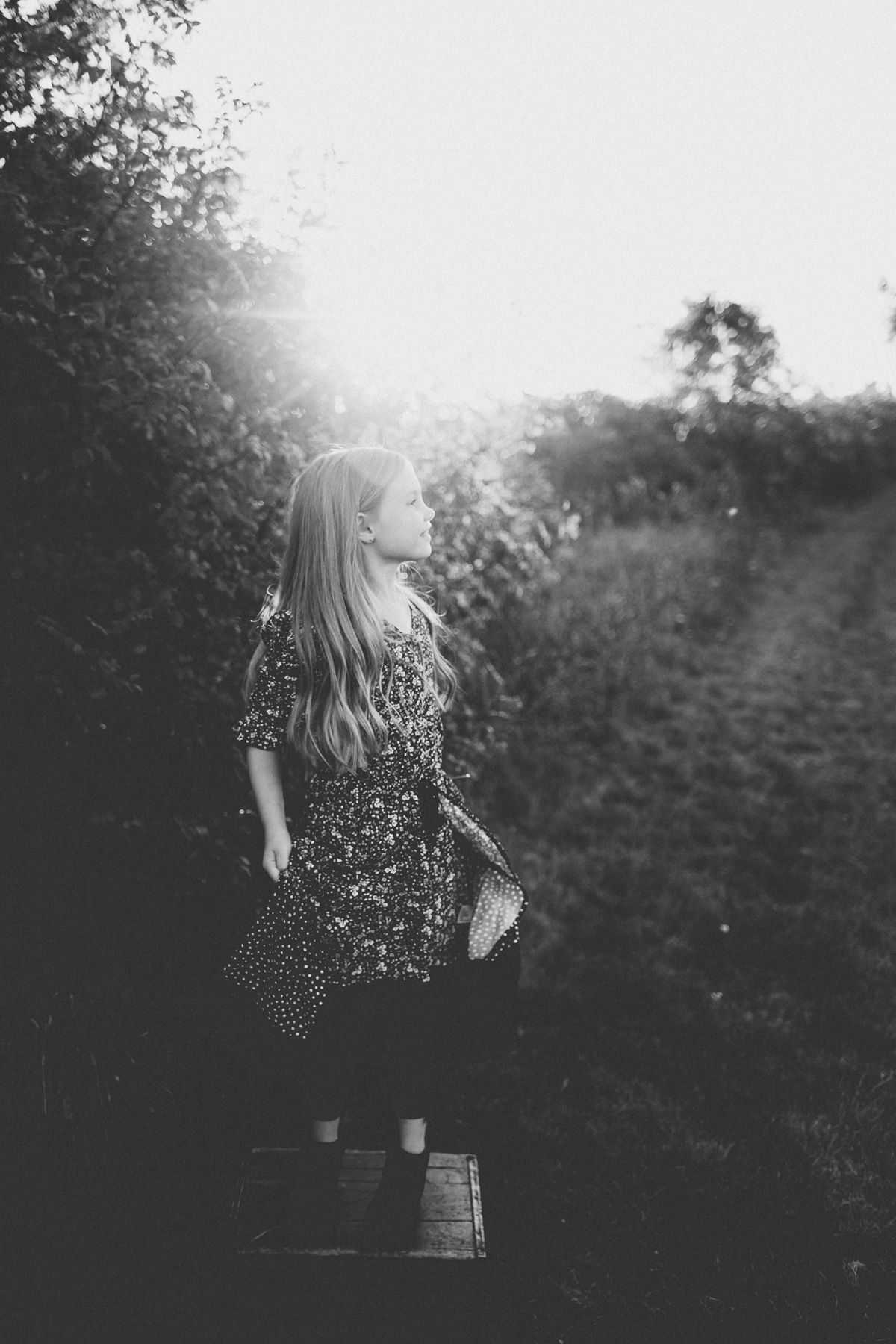 A dreamy black and white portrait of someone in a floral dress standing in a sunlit garden path.