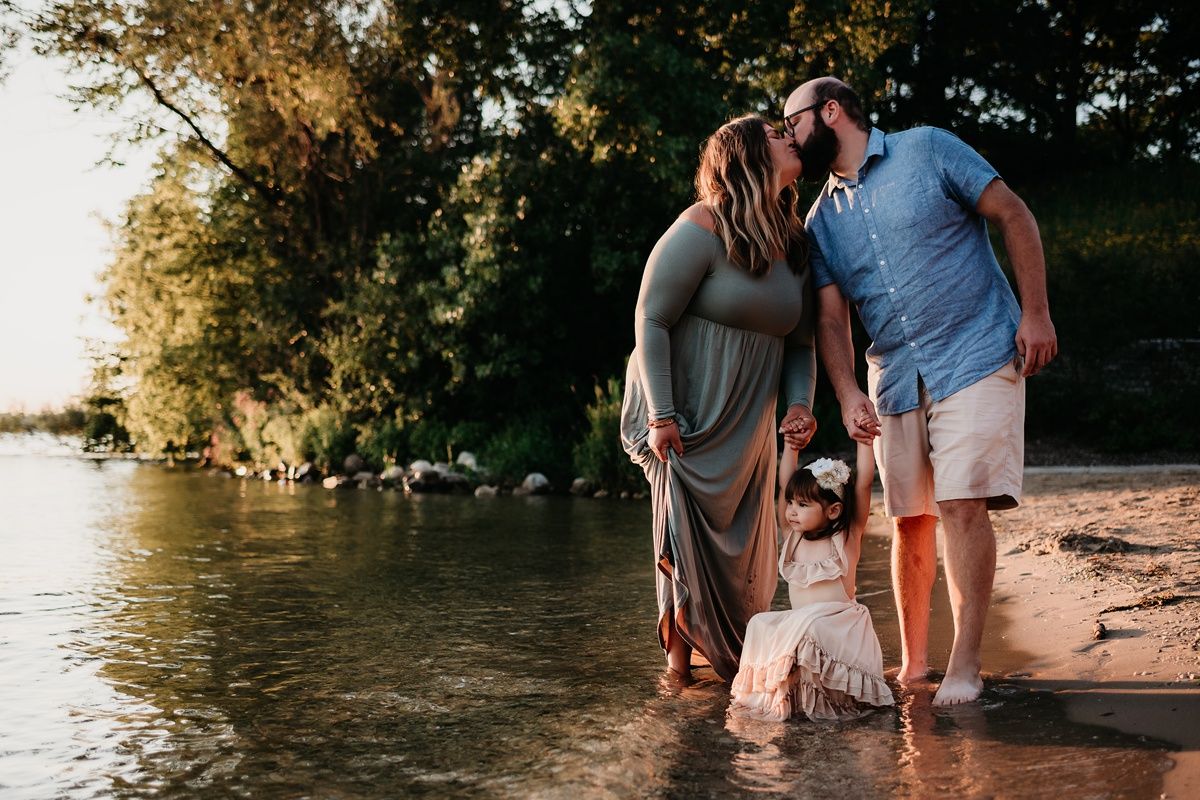 Family wading together in shallow water at sunset near wooded shoreline.