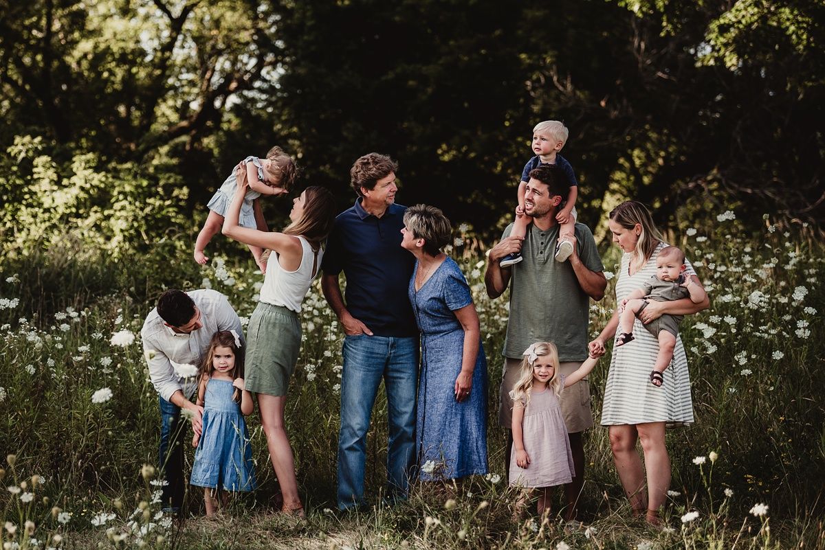 Extended family gathering in a sunny meadow filled with white wildflowers.