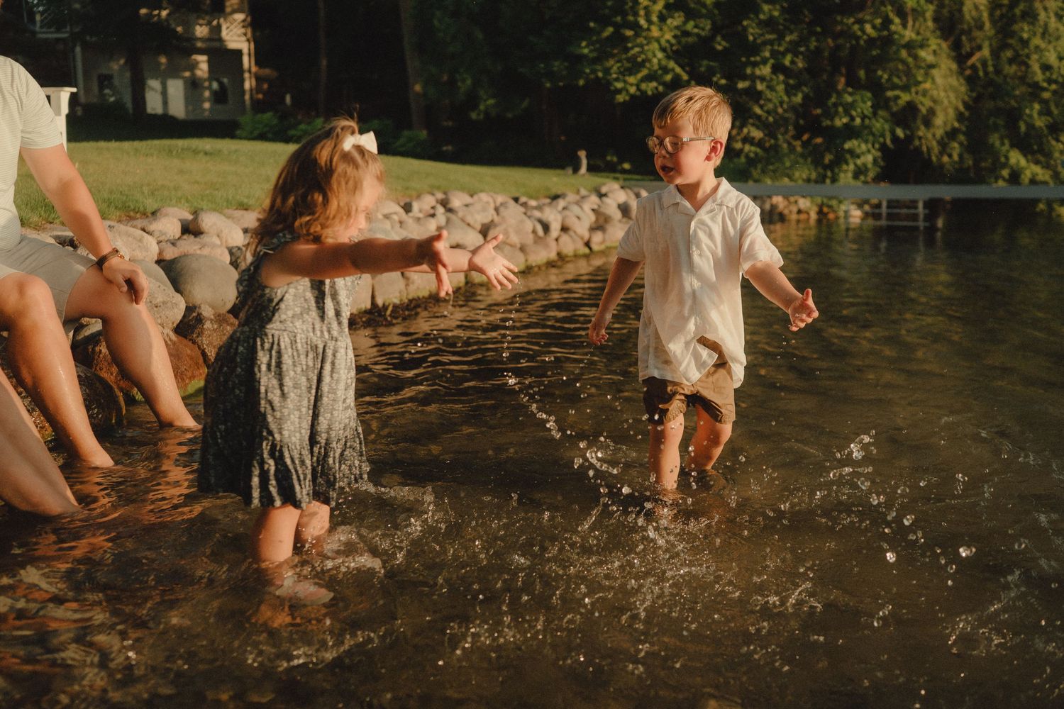 Two children splashing and playing in shallow lake water near rocky shore in warm golden sunlight.