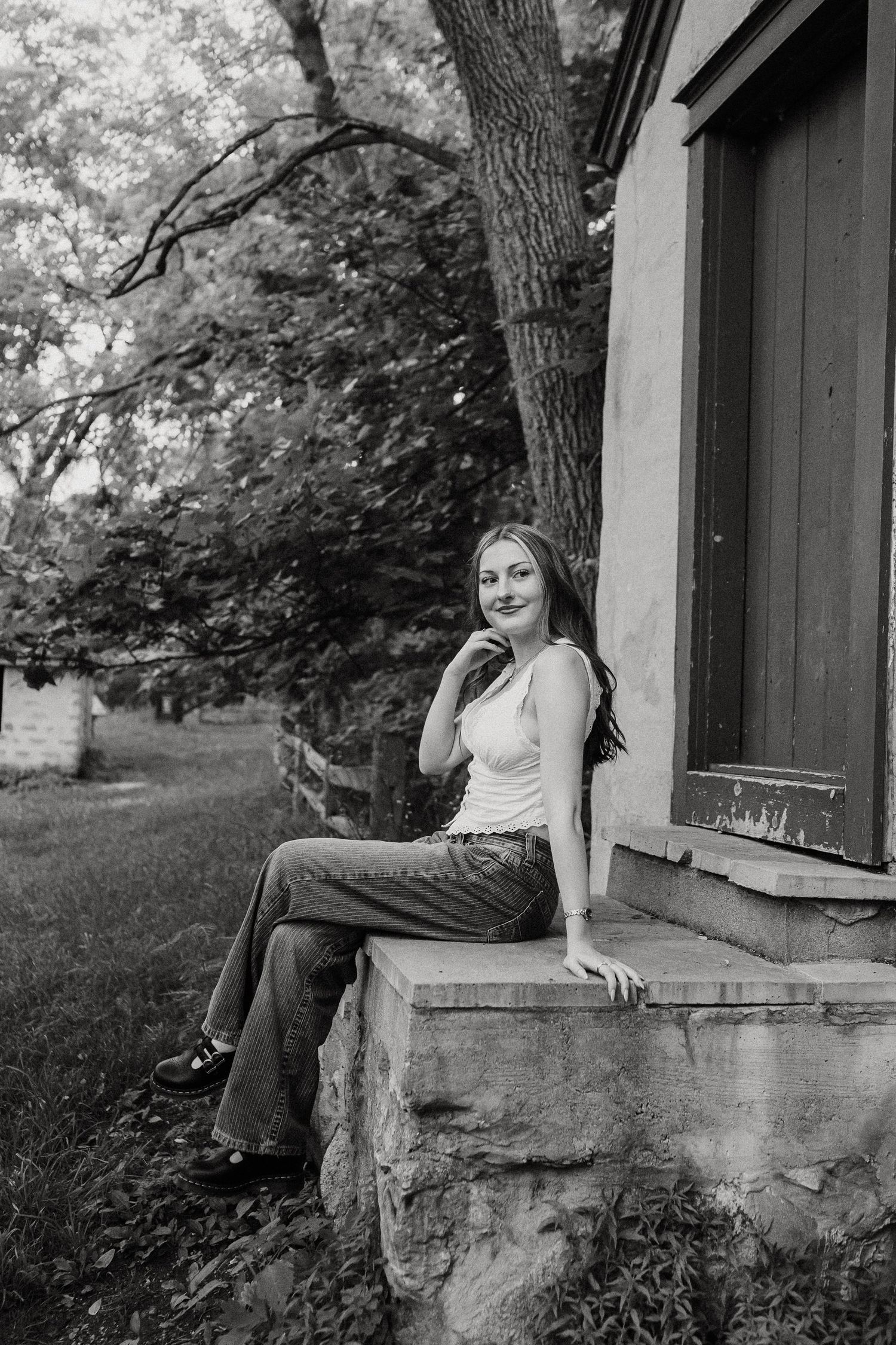 Black and white portrait of young woman sitting on stone ledge beside vintage building outdoors.