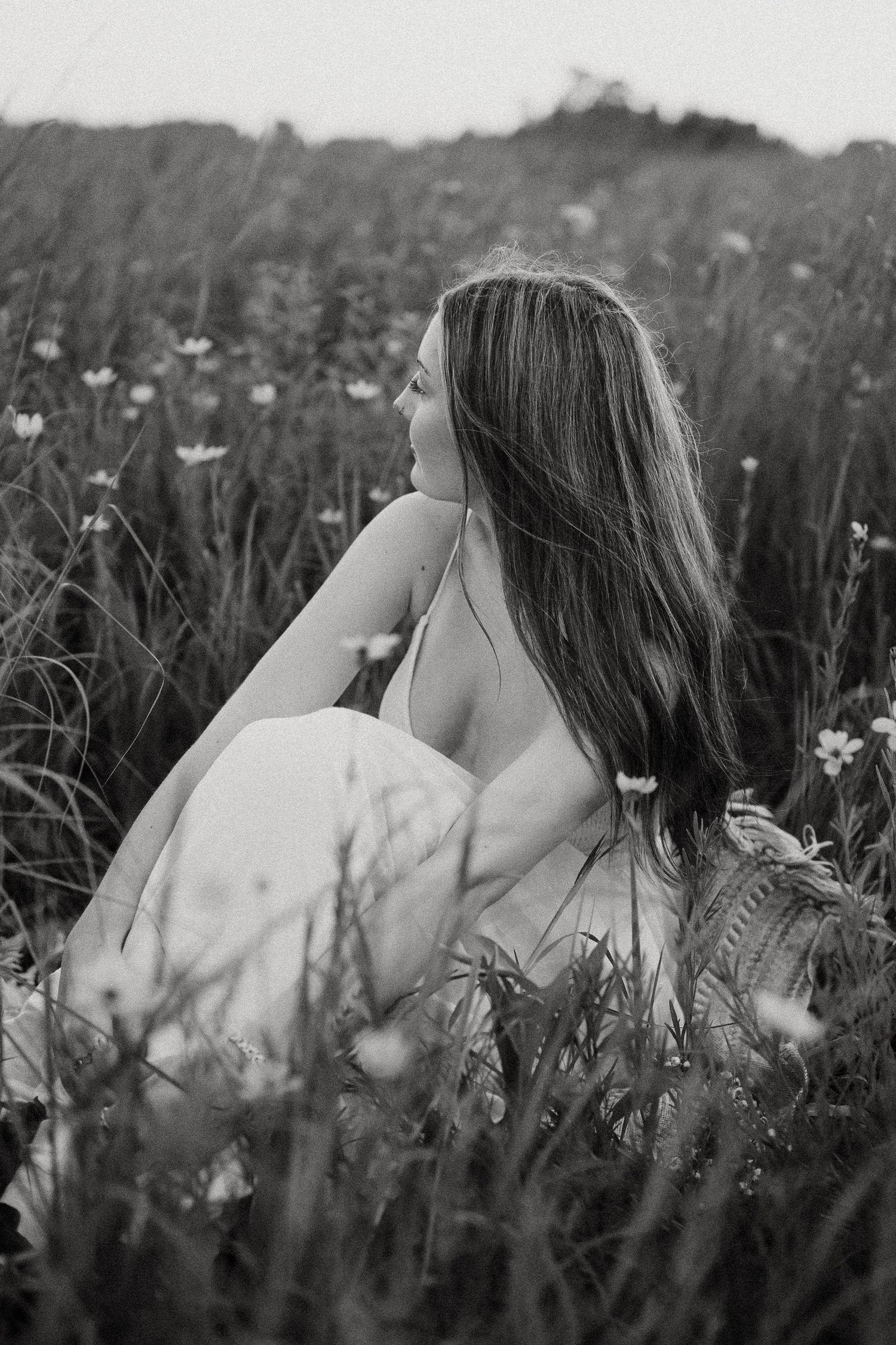 Black and white photo of a young woman sitting in a wildflower field, gazing into the distance.