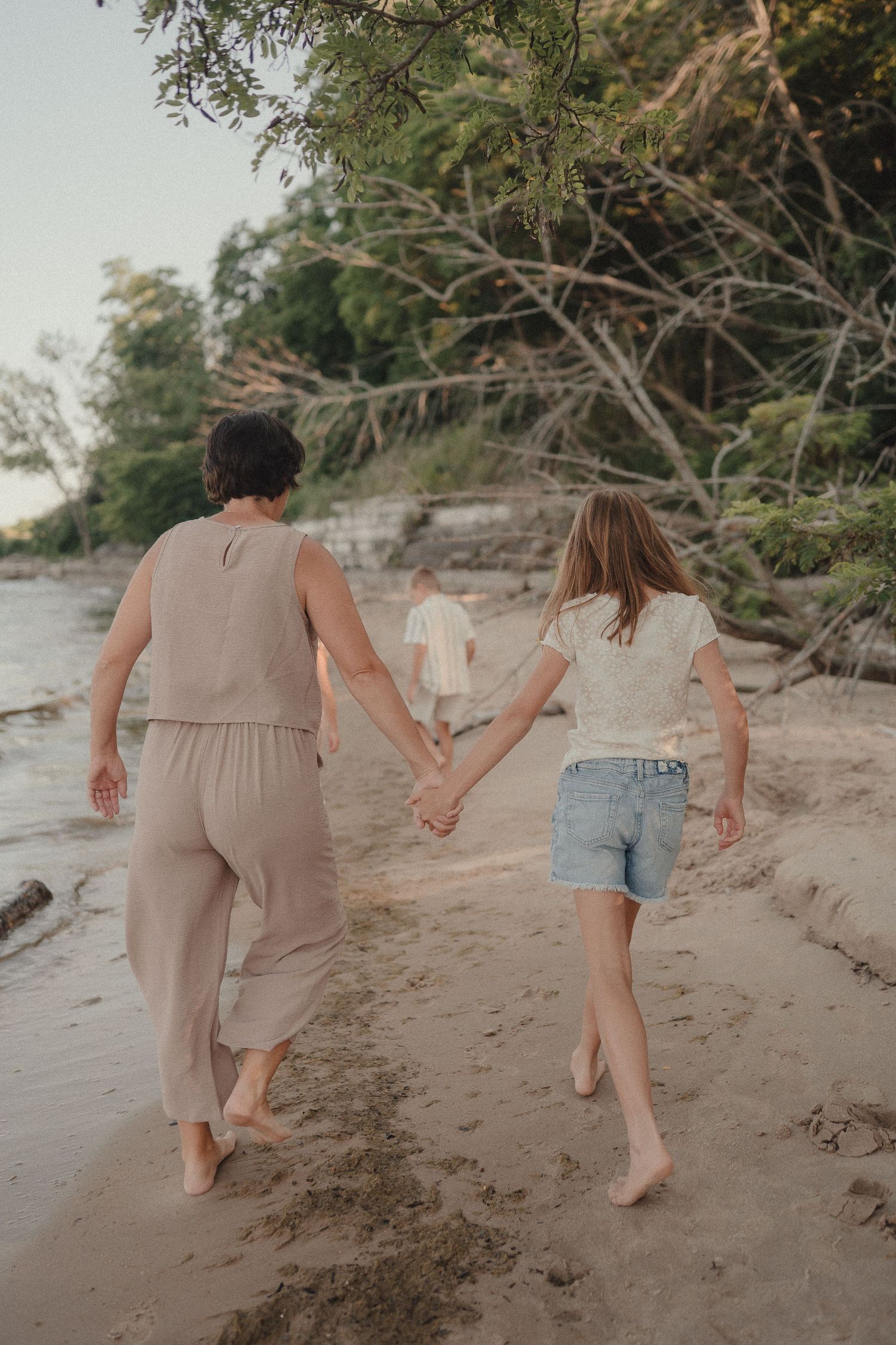 Mother and daughter hold hands walking barefoot on sandy beach with trees in background.