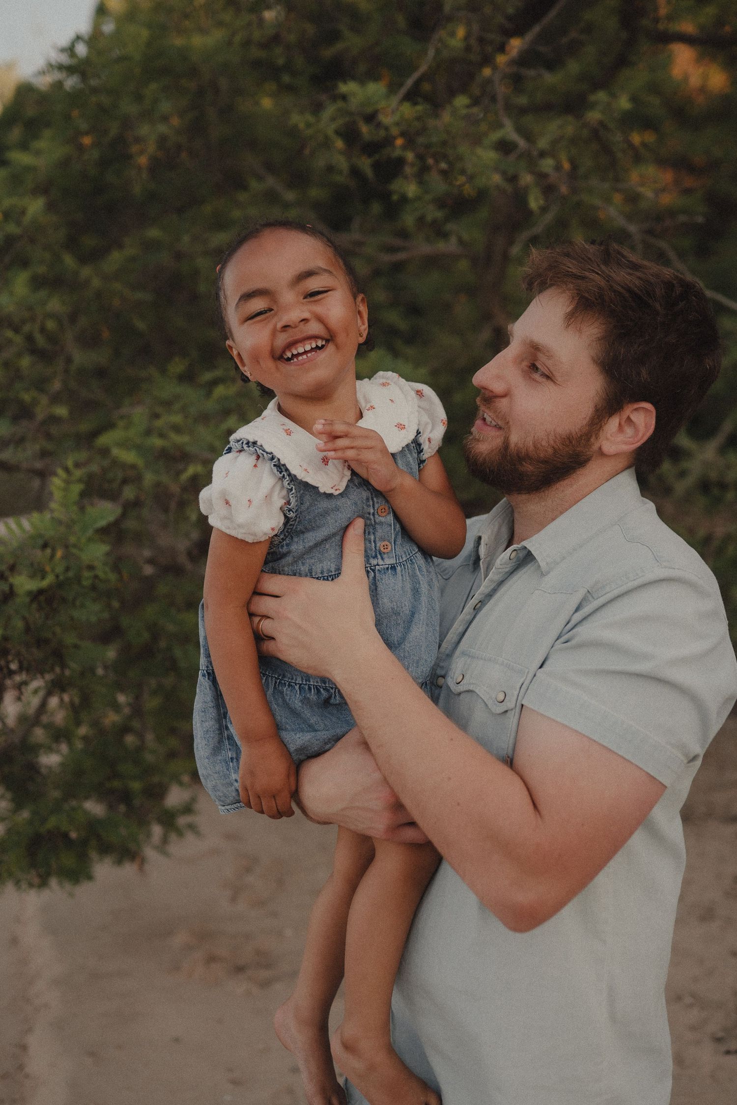 A bearded man holds a laughing young girl in a denim dress outdoors on a sandy beach with green trees.