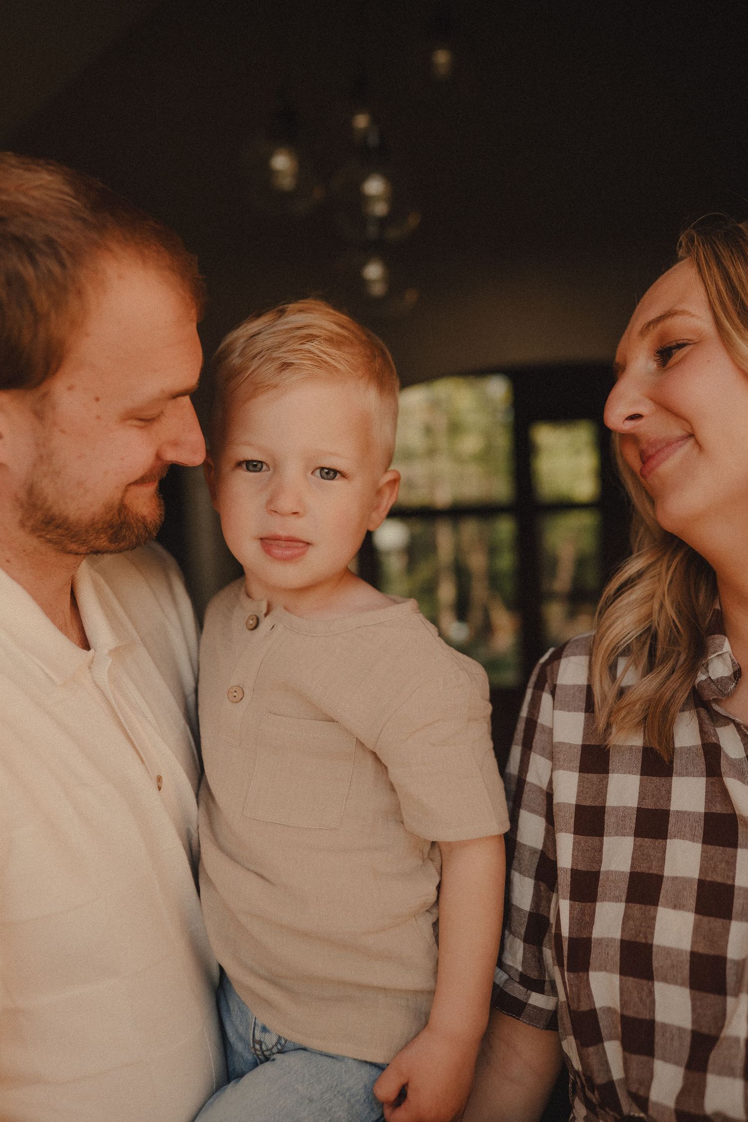 A young blonde toddler stands between his smiling parents indoors in warm golden light.